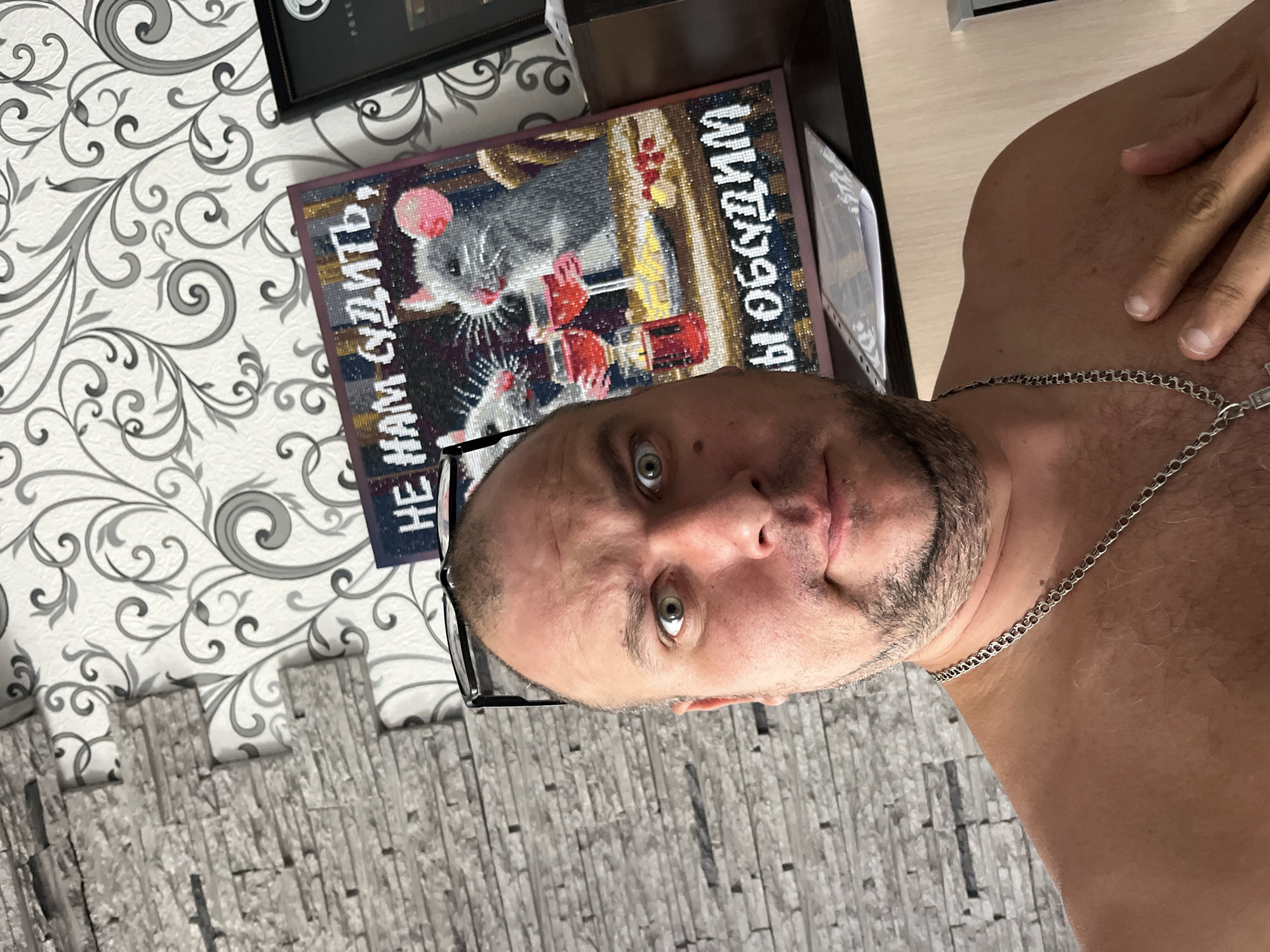 A man is taking a selfie in front of a wall with patterned wallpaper. He is shirtless and wearing a silver chain. Behind him, to the right, is a framed artwork of two mice with wine. The man's glasses are perched on his head. The background wall has a textured stone pattern on the left and floral wallpaper on the right.