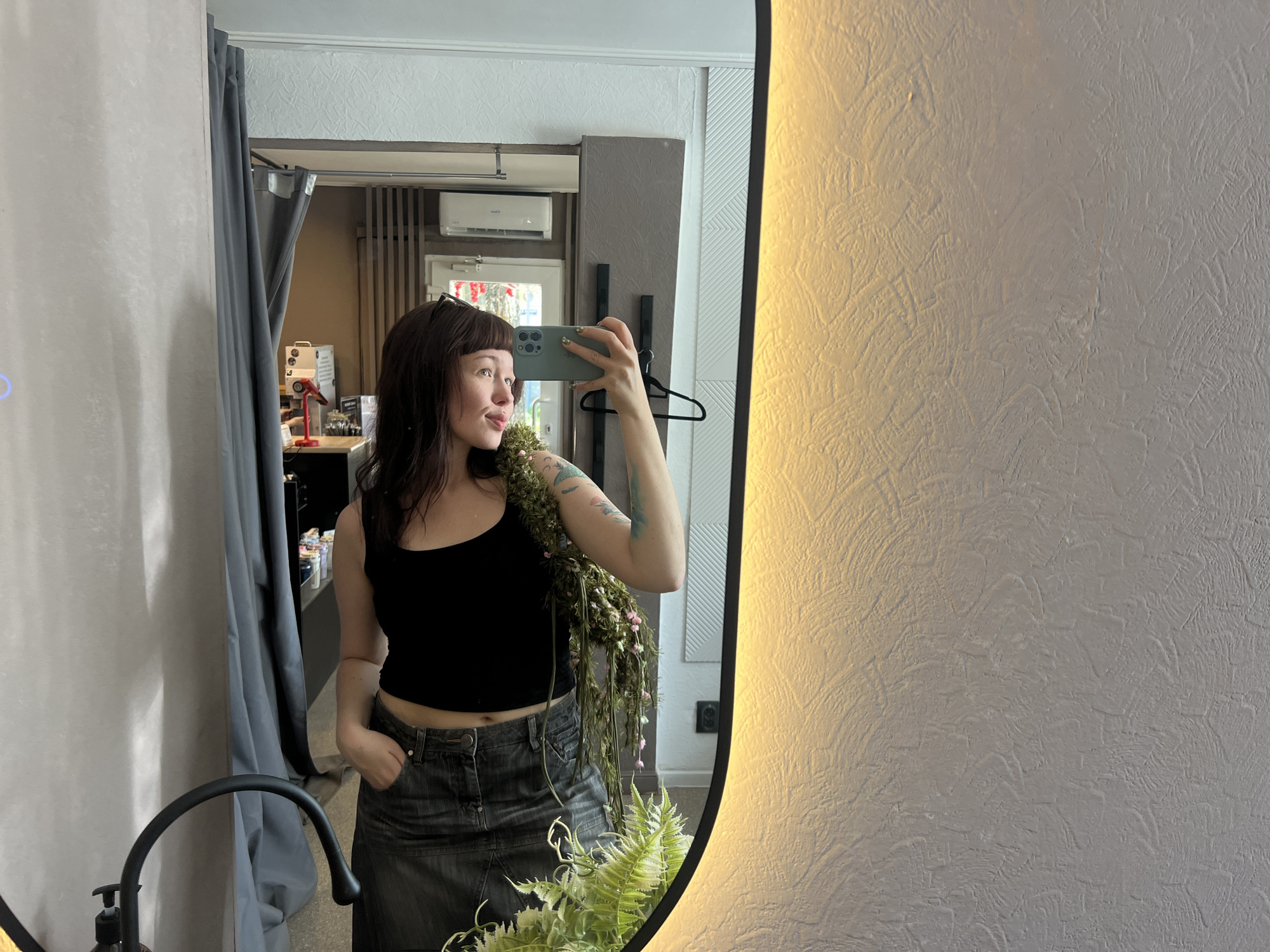 The image shows a young woman taking a selfie in a mirror. She is wearing a black tank top and a denim skirt. She has dark hair and is holding a smartphone in her right hand. A green, floral-like garland is draped over her left shoulder. In the reflection of the mirror, a room is visible with a curtain, an air conditioner unit, and some shelves with various items. The lighting in the room appears to be warm and directional, casting a glow on the textured wall to the right of the mirror. There are also plants visible in the foreground, both in the reflection and potentially outside the frame. The location is identified as Chaikovsky, Russia.