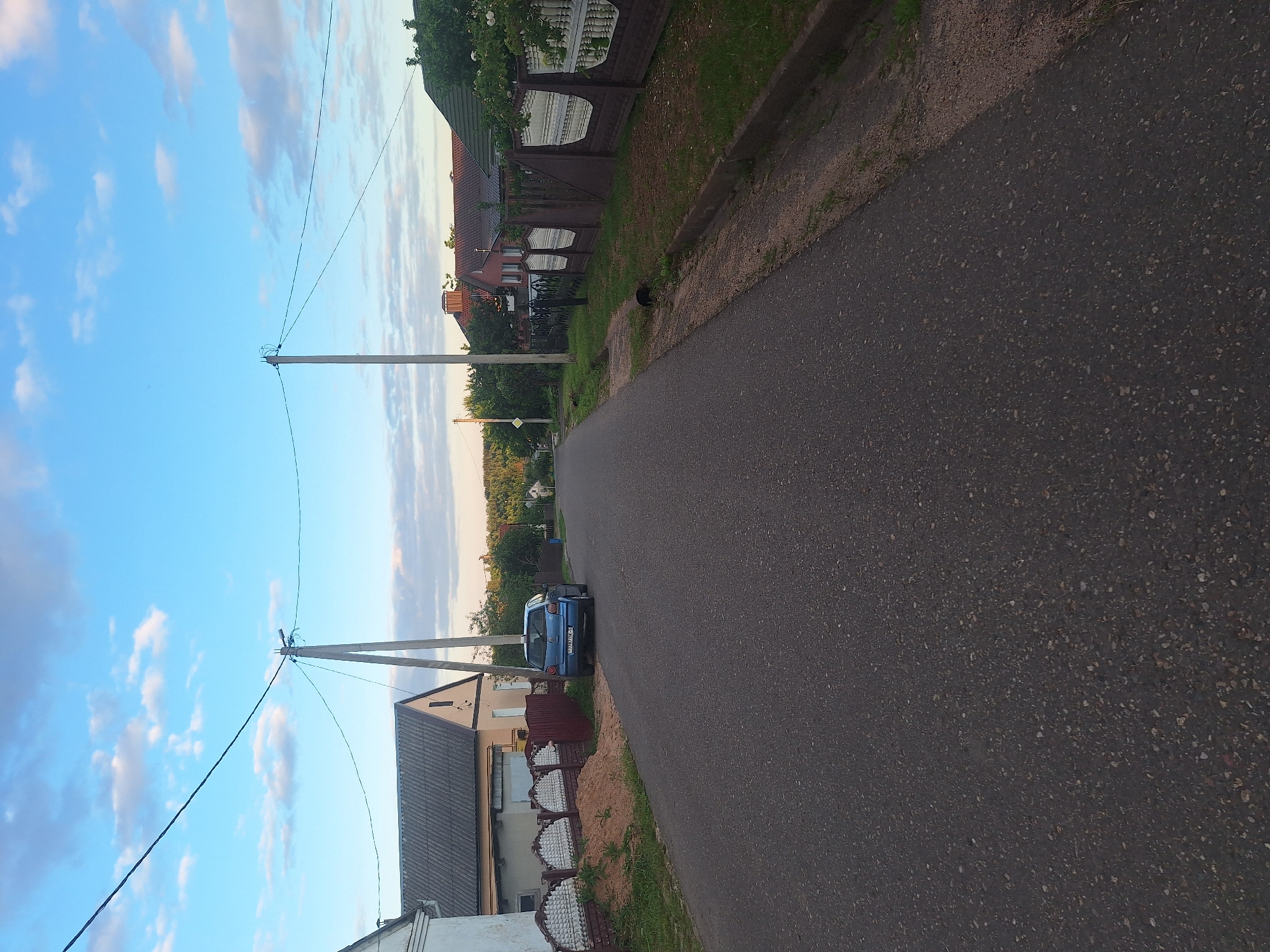 The image shows a street scene in a rural area, likely in the Капыльскі сельскі Савет, Belarus. The asphalt road dominates the foreground, leading into the distance. On the left side of the road, a light-colored house with a grey roof is visible, bordered by a decorative fence with brick and white elements and a pile of sand. A dark blue car is parked on the side of the road in front of a wooden fence. Power lines are strung across the frame, supported by two utility poles. On the right side of the road, a brown fence with decorative white elements separates the road from a grassy area. In the background, several houses with red roofs are nestled among trees, and the sky is a mix of blue with scattered white clouds, suggesting it is late afternoon or early evening. The environment appears residential and quiet.
