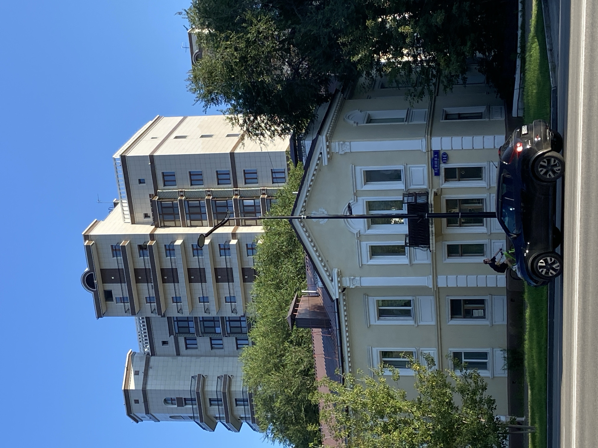 A modern, multi-story apartment building with beige and brown accents stands behind a smaller, cream-colored building with classical architectural details. Both buildings are set against a clear blue sky. In the foreground, a black car is parked on the street, and a person is riding a bicycle on a sidewalk next to a grassy median. Trees line the street, providing shade and a natural element to the urban landscape. A street sign that reads "ЛЕНИНА 114" (Lenina 114) is visible on the side of the cream-colored building, indicating the location as Lenina Street, number 114, in Blagoveshchensk, Russia.