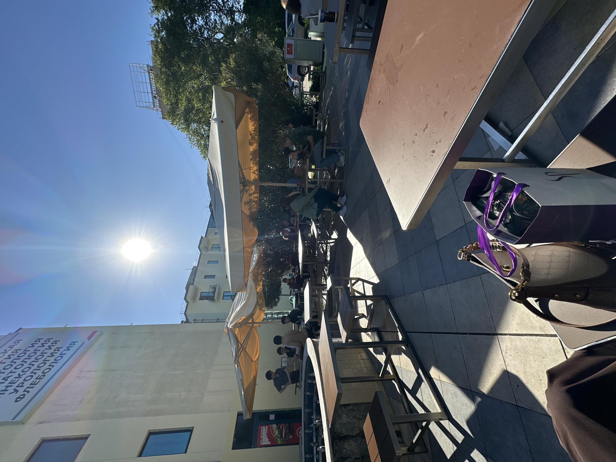 This image, taken in Sochi, Russia, captures a vibrant outdoor cafe scene on a bright, sunny day. The perspective is angled, looking across a paved patio area equipped with numerous tables and chairs. The sun, high in the clear blue sky, casts strong highlights and distinct, long shadows from the furniture and large, light-colored umbrellas, indicating either a late morning or early afternoon.
Several people are visible seated at the cafe tables. Some are comfortably shaded by the umbrellas, while others are in direct sunlight. They appear to be patrons casually enjoying the outdoor ambiance, possibly socializing or dining. In the immediate foreground, a brown cafe table is prominent, and on the ground next to it, a purple shopping bag with white handles and a handbag are visible.
The urban setting includes buildings and lush green trees in the background. A large sign on a building displays Cyrillic text, clearly indicating medical specialties such as "МАТОЛОГИЯ" (likely part of Dermatology), "ГИНЕКОЛОГИЯ" (Gynecology), "УРОЛОГИЯ" (Urology), and "ФЛЕБОЛОГИЯ" (Phlebology), suggesting the proximity of a medical facility. Below this, a smaller red sign partially reveals the word "БУРГЕР" (Burger), possibly pointing to a nearby fast-food restaurant. Above the trees and buildings, a metal framework of an empty billboard structure is visible against the bright sky, with the sun itself causing a significant lens flare. The overall impression is one of a relaxed, bustling outdoor space on a pleasant day.