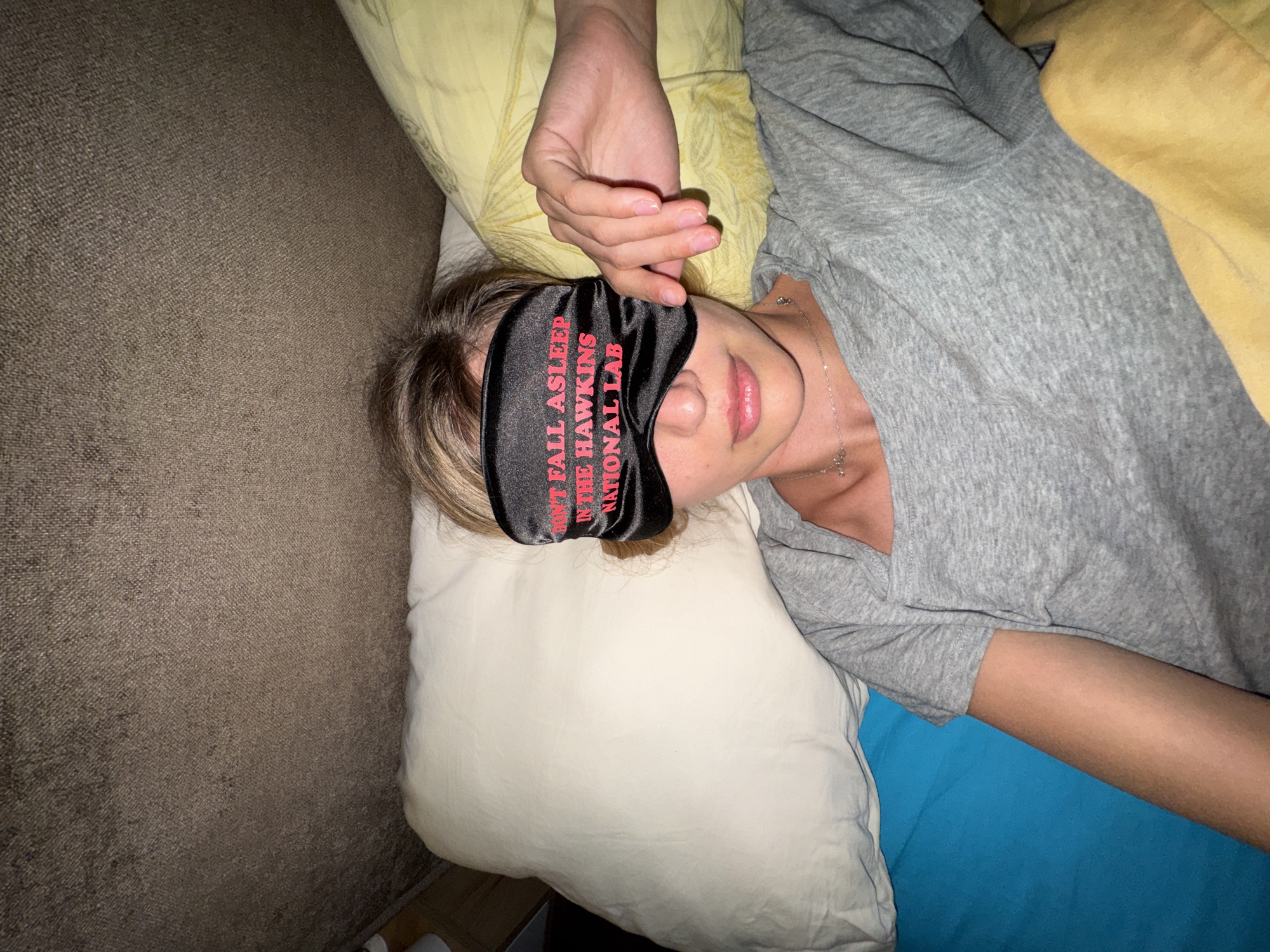 A person lies on a bed, wearing a sleep mask with the words "DONT FALL ASLEEP IN THE HAWKINS NATIONAL LAB". The individual is positioned on a white pillow, with a grey shirt and blue bedding visible. A tan couch is seen adjacent to the bed. Location is Unknown, Unknown.