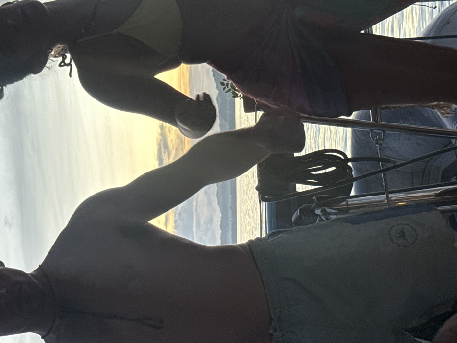 The image depicts two individuals on a boat at either sunrise or sunset, silhouetted against a bright sky over a body of water, likely in the coastal area of General Luna, Philippines. The left individual, appearing male, is shirtless and wears light-colored shorts with a circular logo on the left thigh, and a dark cord necklace. The right individual, appearing female, wears a light-colored bikini top and a darker, patterned lower garment, with hair styled upwards. Their upper bodies are visible, with their arms extended towards each other, suggesting proximity or an interaction, possibly holding hands, though their hands are partially obscured. Below them, visible boat structures include metal railings and a coiled dark rope. In the background, a calm body of water extends to distant landmasses with low-lying hills or mountains, beneath a sky displaying hues of orange, yellow, and light blue. A small potted plant is visible on the right side of the boat.