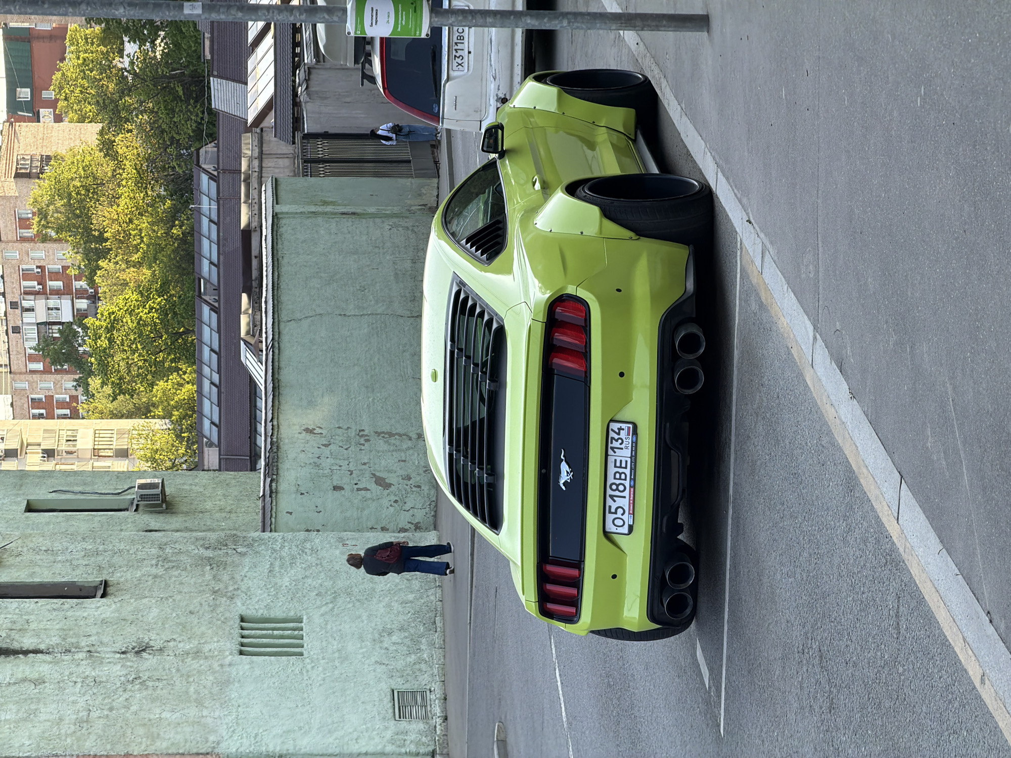 A lime-green Mustang is parked on a city street in Moscow, Russia. The license plate number is "O518BE 134 RUS." The car is parked in a designated parking space, and the back of a white SUV is visible in front of it. A person wearing blue jeans and a maroon jacket is walking along the sidewalk near the green wall. A building with an air conditioner and various windows and vents is next to the sidewalk and wall. Other brick buildings with green trees nearby are also in the image.