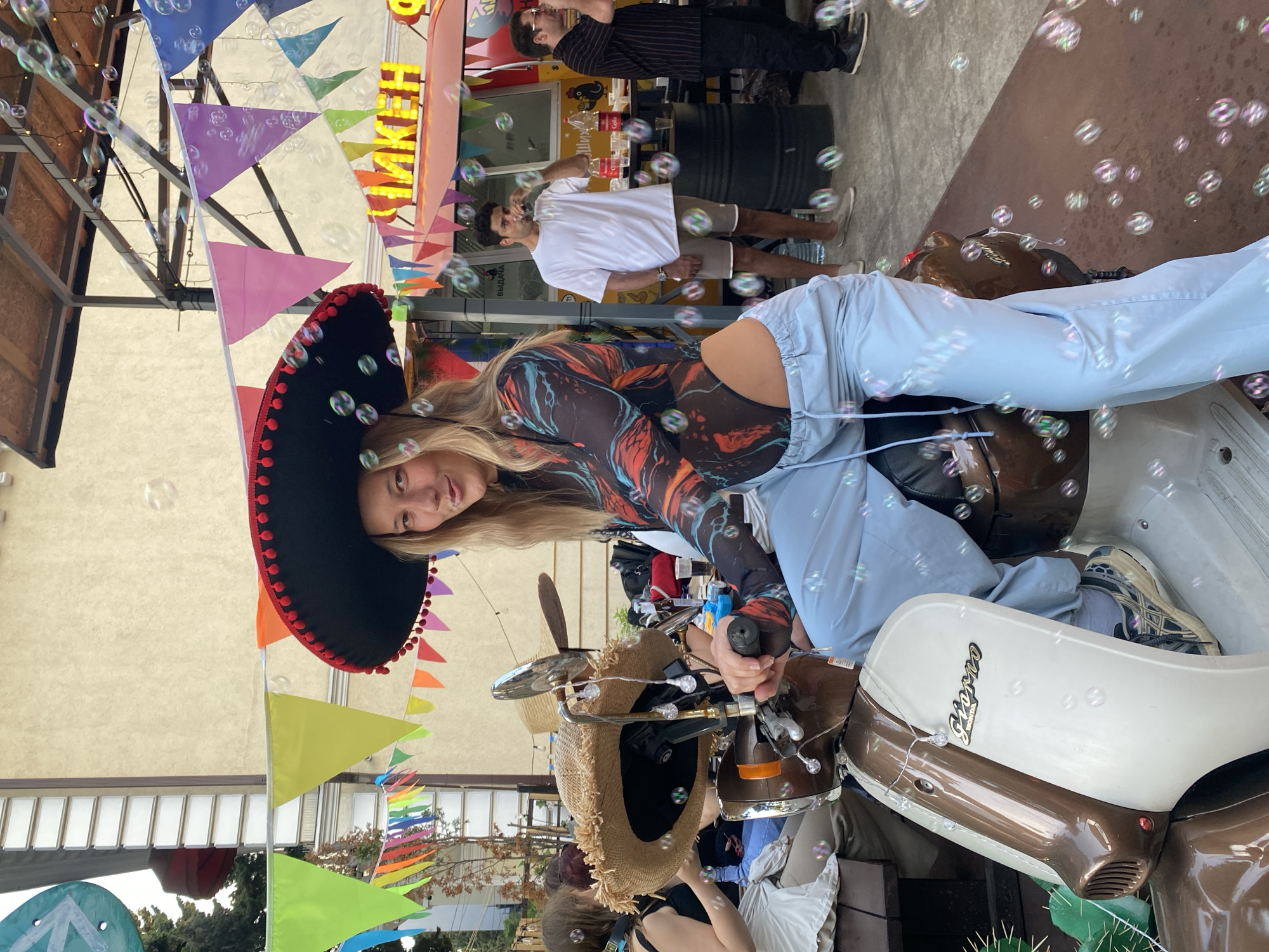 The image depicts an outdoor festive scene, identified as being in Sochi, Russia, due to the presence of Cyrillic signage. The central subject is a young adult female with long blonde hair, wearing a large black sombrero with a red pom-pom trim. She is positioned on a two-tone brown and off-white Honda Giorno scooter, which is adorned with a string of decorative lights and two straw hats draped over its handlebars. The woman wears a multi-colored abstract print bodysuit (featuring blues, oranges, and black) and light blue, drawstring-waist track pants pulled low. She faces the camera with a playful expression, her tongue slightly protruding.
Numerous transparent soap bubbles are floating throughout the entire frame, from the foreground to the background, creating a whimsical atmosphere. Above the scene, colorful triangular bunting in various hues (red, orange, yellow, green, light blue, pink, purple) is strung horizontally.
In the mid-ground to the right, a red food truck or stall is visible, featuring illuminated yellow Cyrillic letters spelling "ЯКЕН" and smaller white Cyrillic text reading "ВЫДАЧА" (meaning "Issuance" or "Pickup"). A man with dark hair, wearing a white t-shirt, beige shorts, and light slip-on shoes, stands near the food truck, drinking from a dark glass. To his right, a second man in a striped shirt is partially visible. On the left mid-ground, several individuals are seated at a wooden table, mostly obscured by the scooter. A light-colored building is visible in the background, and trees line the left side of the frame. The ground appears to be a paved surface. In the bottom left, a green artificial plant resembling a cactus is partially visible.