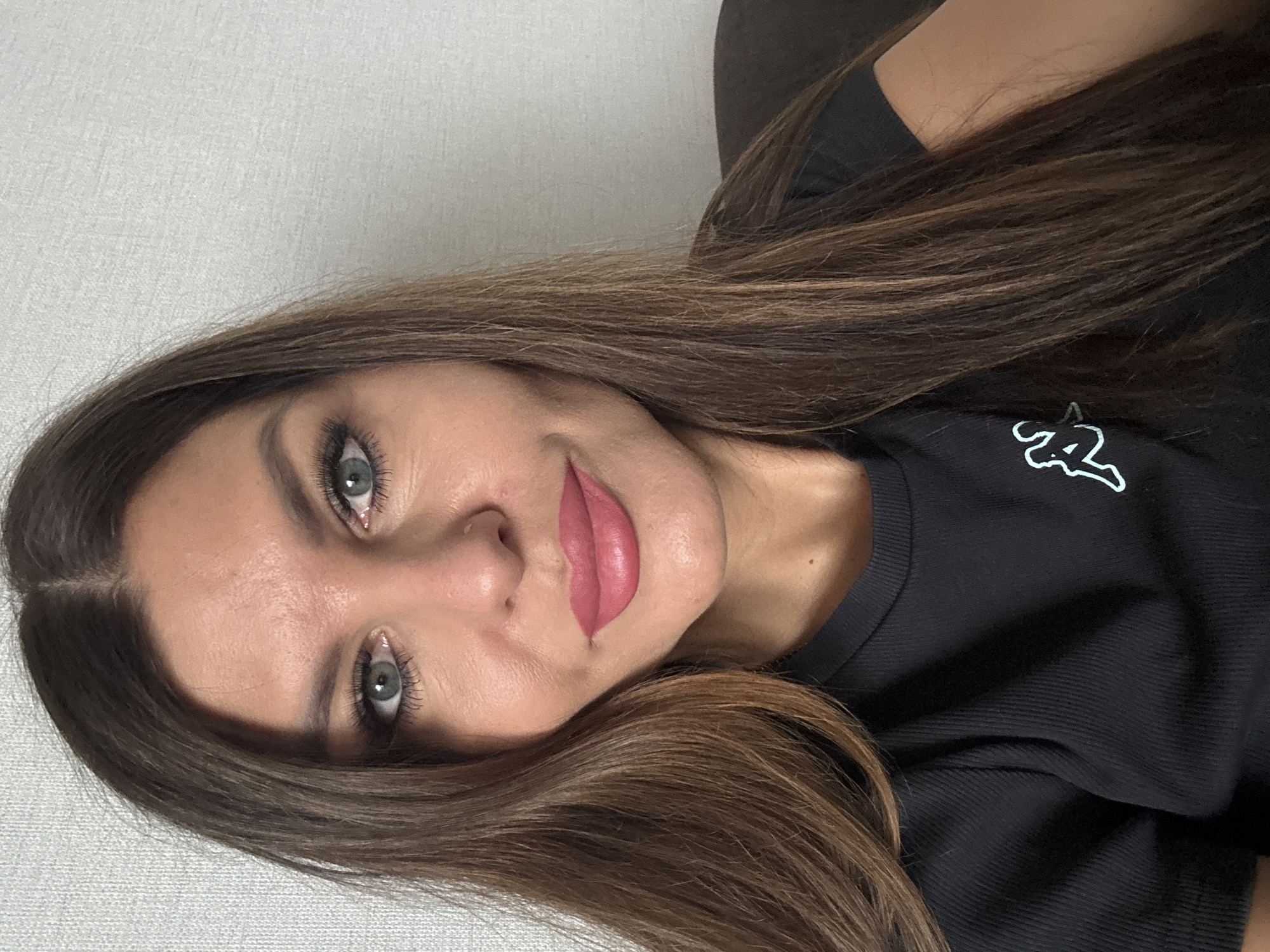 A woman with long, brown hair is wearing a black t-shirt with a white logo. Her eyes are gray, and she is wearing pink lipstick. The background appears to be a plain, light-colored wall. The image does not show any evidence of Chaikovsky, Russia.