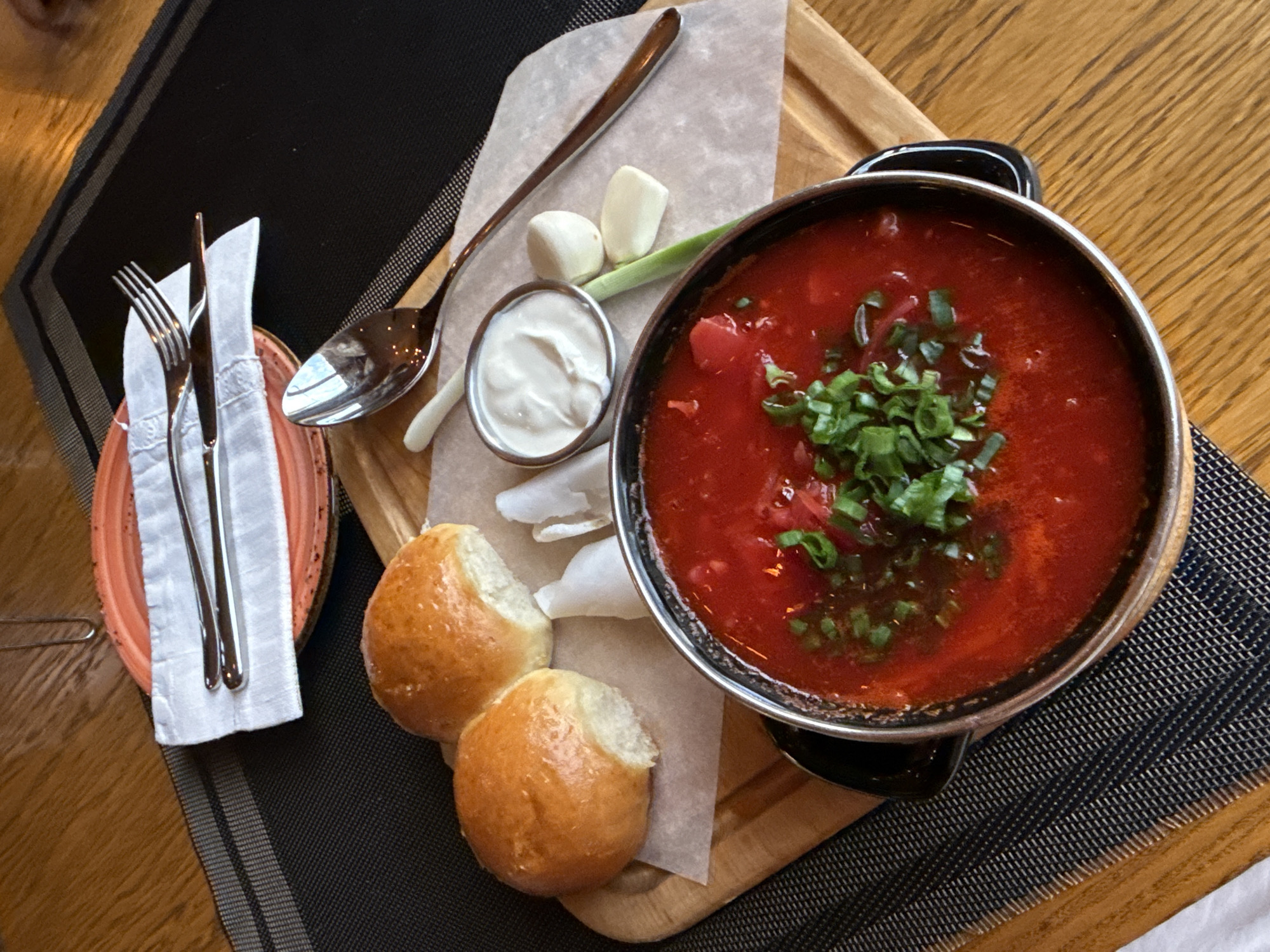 A close-up, overhead shot showcases a rustic dining scene, likely in Olginka, Russia, featuring a bowl of vibrant red borscht garnished with chopped green onions. To the left of the soup, two golden-brown bread rolls sit on a piece of parchment paper, nestled on a wooden board. Beside the borscht, a small metal ramekin holds a dollop of white sour cream, accompanied by three cloves of peeled garlic and a green onion stalk. A large, reflective spoon rests near the sour cream, and another set of silverware, a fork and a knife, are neatly arranged on a folded white napkin atop a coral-colored plate. The wooden table has a visible grain, and a dark placemat with thin gray stripes provides a base for the meal. The overall impression is one of a hearty, traditional meal served in a comfortable setting.