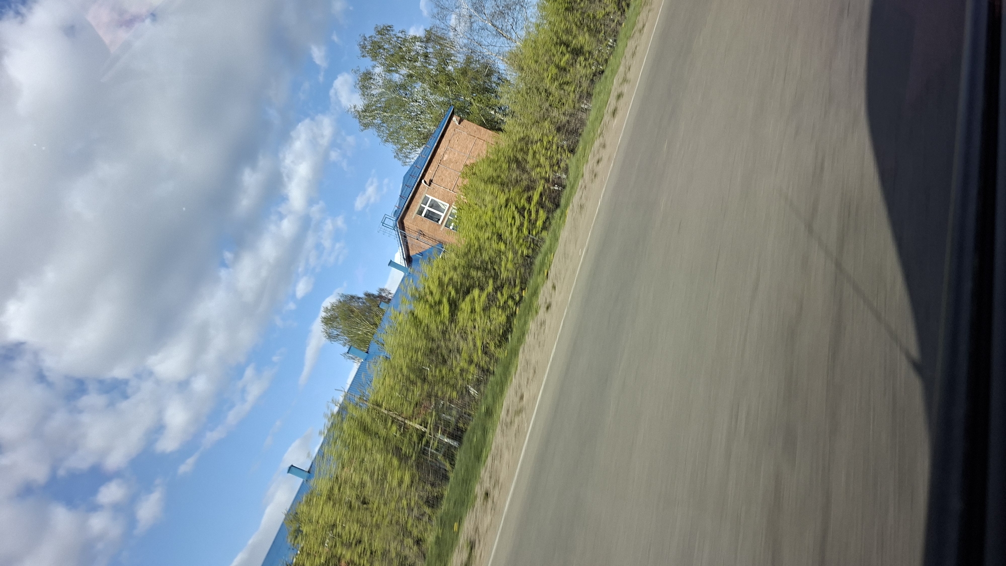 The image shows a road next to a blue fence with greenery. Beyond the fence is a brick building with a blue roof. In the background, there is a blue sky with scattered clouds. The photo was taken from a moving vehicle. Based on the presence of the brick building and the blue fence, this location may be found in the city of Chaikovsky, Russia.