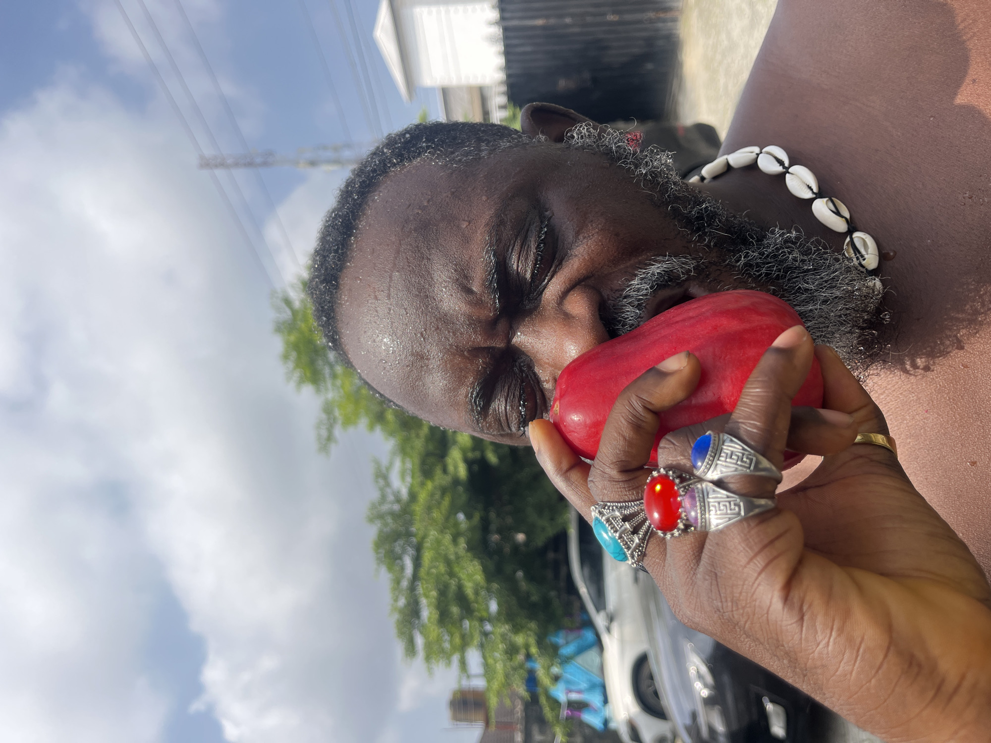 A close-up, low-angle shot captures a dark-skinned man in Ado, Nigeria, his face partially obscured by a bright red fruit he is holding to his nose. He has a graying beard and is sweating. He wears several rings on his left hand, including one with a large red stone and another with a turquoise stone. A necklace made of white cowrie shells is visible around his neck. In the background, trees, a fence, a white building, and parked cars are visible under a bright, cloudy sky. The overall impression is one of an outdoor, possibly humid, environment.