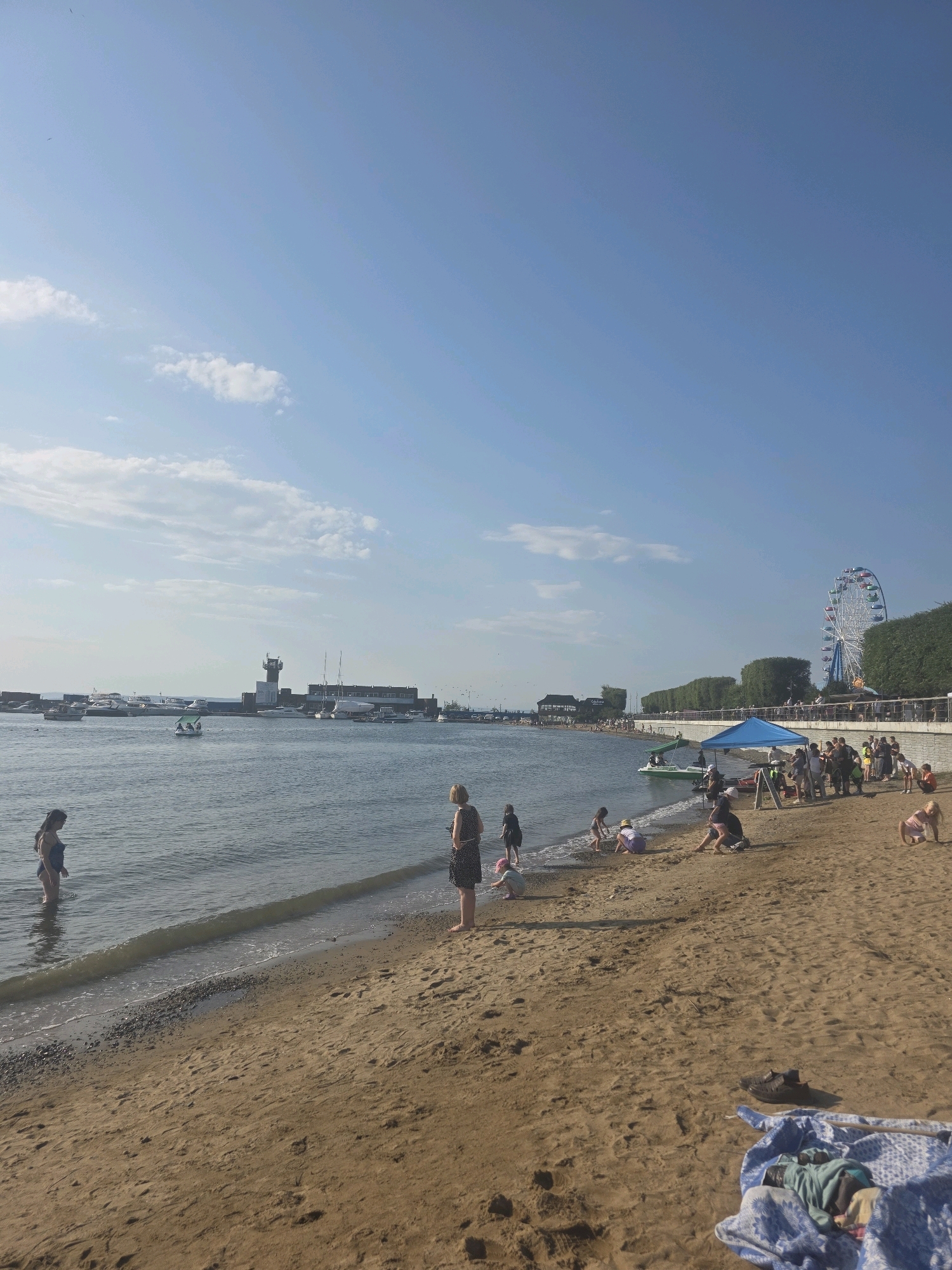 The image depicts a sunny day at a beach, likely in Vladivostok, Russia, characterized by a clear blue sky with scattered clouds. In the foreground, a sandy beach is populated by various individuals, including children playing in the sand and at the water's edge. Some adults are visible standing and observing. A blue canopy tent is set up on the sand, with several people gathered around it. The water is calm, with a few small boats and a paddleboat visible. In the background, a Ferris wheel stands prominently against the skyline, suggesting it is part of an amusement area or park. A lighthouse and several buildings, possibly port facilities, are also visible along the coastline. Trees line the elevated walkway behind the beach, and more boats are docked in the harbor. The overall scene conveys a leisurely atmosphere, with people enjoying recreational activities at the seaside.
