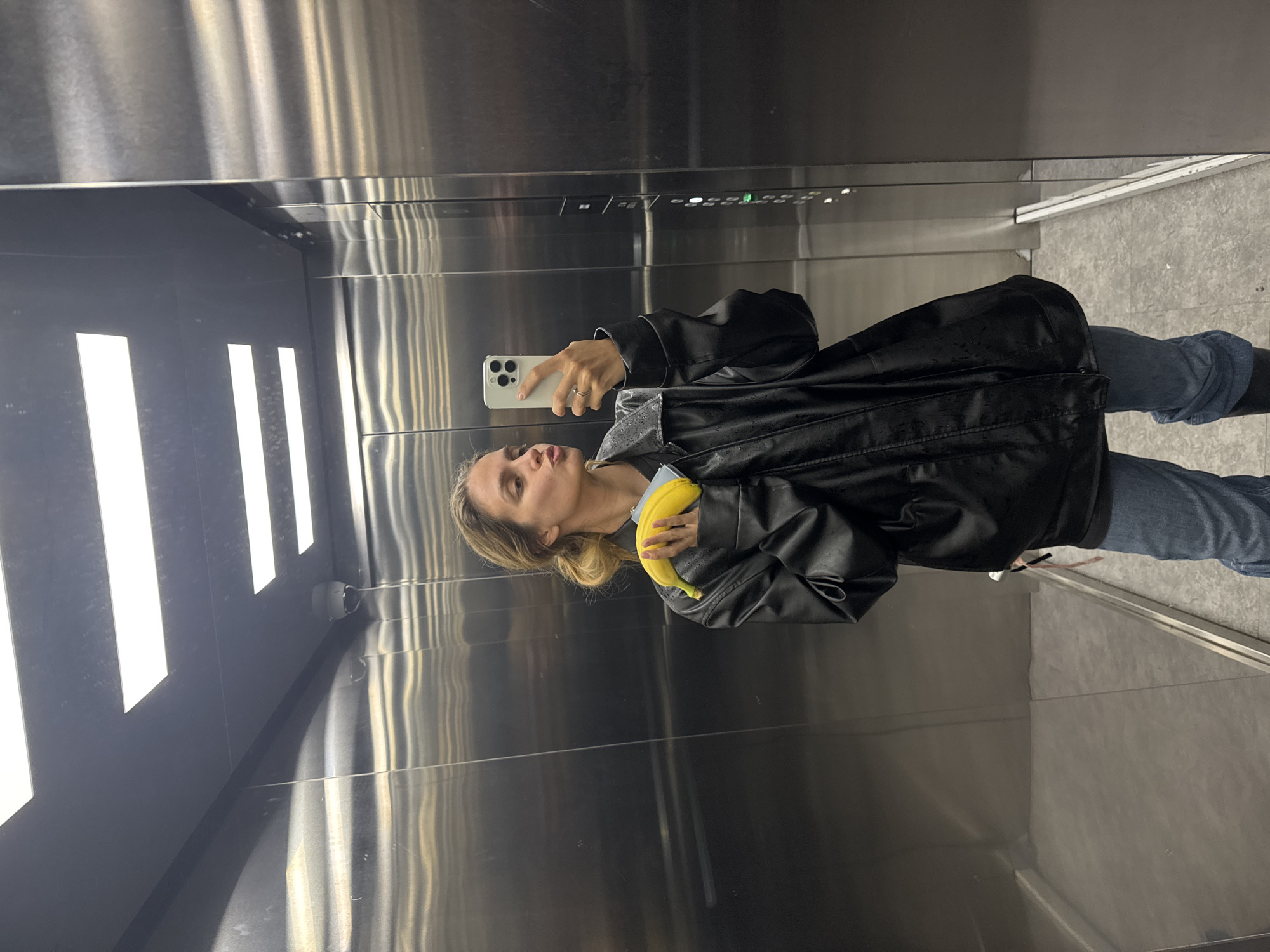 A woman is taking a selfie in what appears to be an elevator. She is holding her phone in one hand and a banana in the other. She is wearing a black leather jacket and jeans. The elevator has brushed metal walls and illuminated vertical light strips. The location is in the city of Unknown, Unknown.