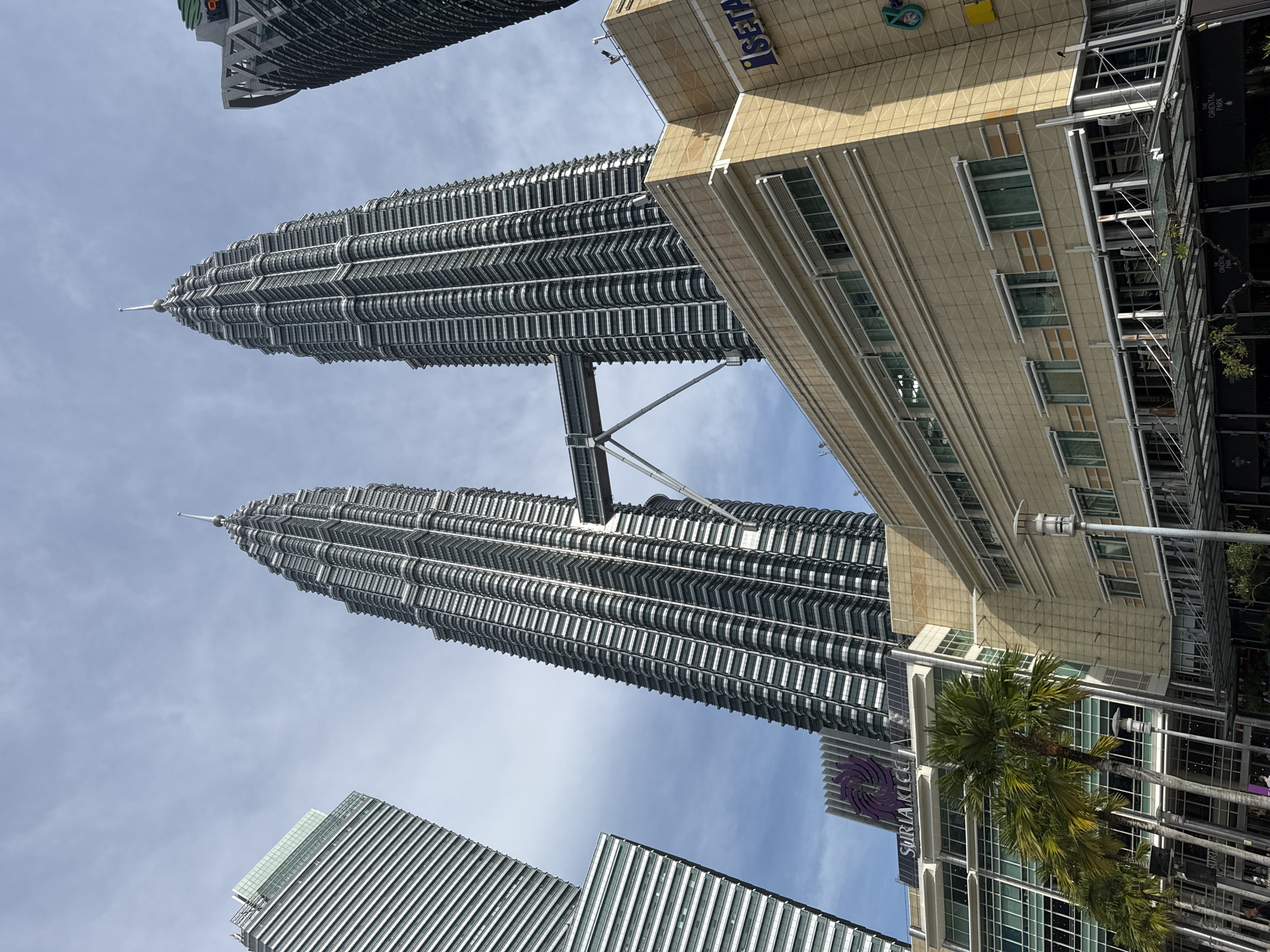 This image presents a low-angle view of the iconic Petronas Twin Towers in Kuala Lumpur, Malaysia, standing tall against a bright blue sky with scattered white clouds. The twin skyscrapers dominate the central frame, showcasing their intricate, modern architecture and the skybridge connecting them at a mid-level. The towers' exteriors are a mix of metallic and glass panels, reflecting the daylight.
In the foreground and to the right, a multi-story building with a light beige facade is visible, displaying the text "ISETAN" in dark blue letters on its upper section. Further down and to the right, lush green palm trees line the street level, partially obscuring another building with a "SURIACLCC" sign and a distinctive purple spiral logo. The text "THE ORIENTAL PARK" is also faintly visible on a banner on one of the surrounding buildings. In the lower left, the tops of other modern buildings with striped, metallic facades are partially visible, contributing to the dense urban landscape. The overall scene captures the grandeur and modernity of Kuala Lumpur's city center on a clear, sunny day.