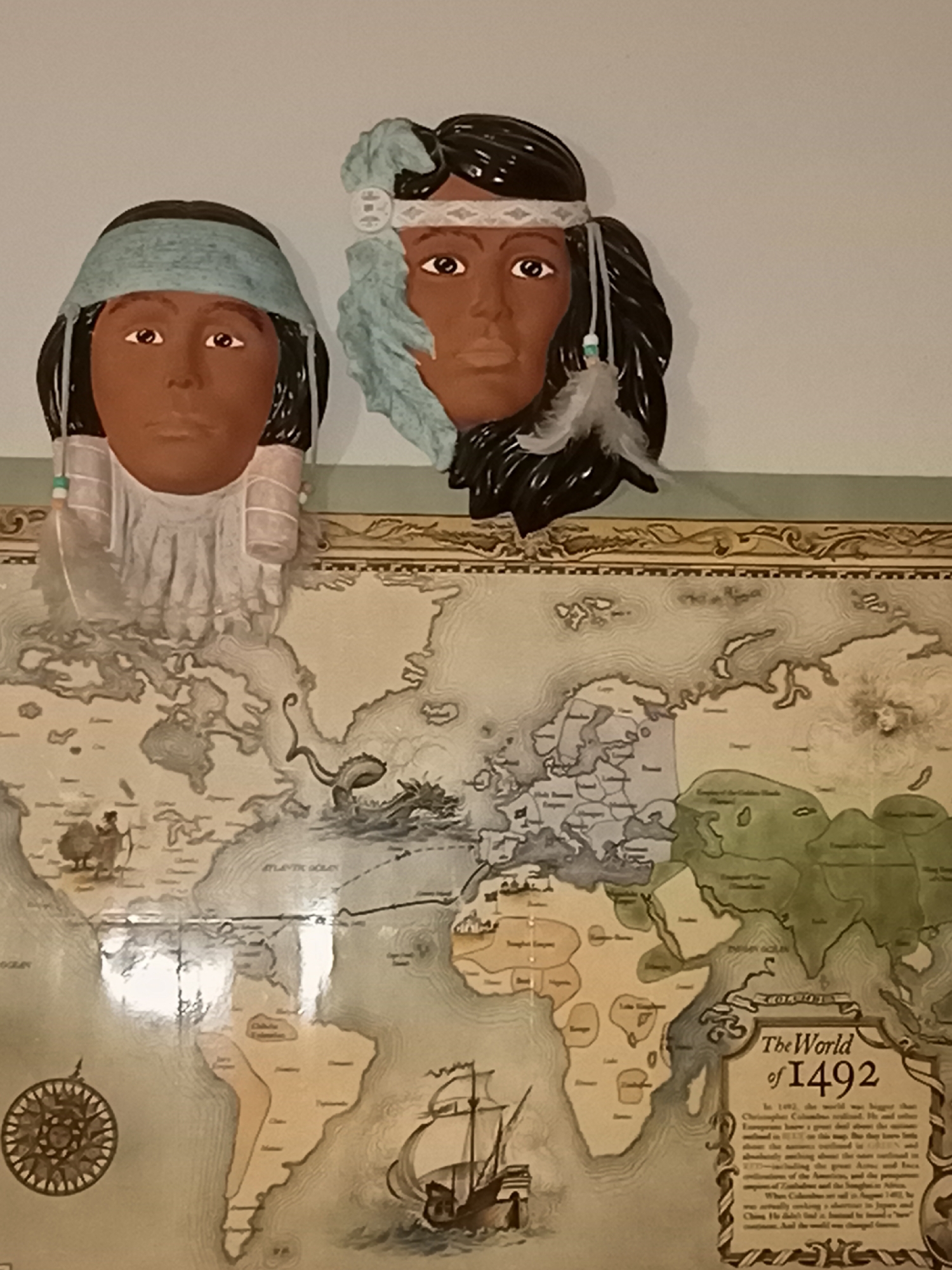 The image shows a wall decoration featuring two Native American-inspired decorative busts above a map. The busts are of young women, each with dark hair and headbands. The one on the left has a light blue headband and white fringe-like decorations around her face. The one on the right has a white headband with a diamond pattern and a light blue fringe around her face. Both have feathers and small beads as adornments.
Below the busts is a decorative world map, titled "The World of 1492." The map is detailed with outlines of continents, oceans, and some political divisions. Various historical scenes and symbols are depicted on the map, including sailing ships and figures of people. A compass rose is visible on the lower left portion of the map, and a text box with historical information is on the lower right. The map and busts appear to be mounted on a wall.
The location is stated to be in the city of Amarillo, United States of America. The environment is an indoor setting, likely a room in a home or public space, with a light-colored wall.