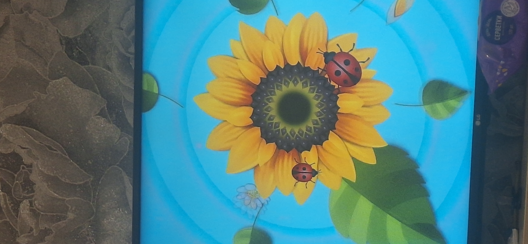 A television screen displays a digital illustration of a sunflower against a bright blue background with radiating circles. Two ladybugs are positioned on the sunflower's petals. To the left of the sunflower, a small, light blue flower is visible. Several green leaves, one large on the left and others smaller and scattered, are also part of the illustration. The bottom of the image shows the black bezel of the television, with an "LG" logo visible on the right side. Below the screen, partially visible, is a purple package of wet wipes labeled "СЕРВЕТКИ" (SERVIETKI). The image captures a television display in Bila Tserkva, Ukraine.