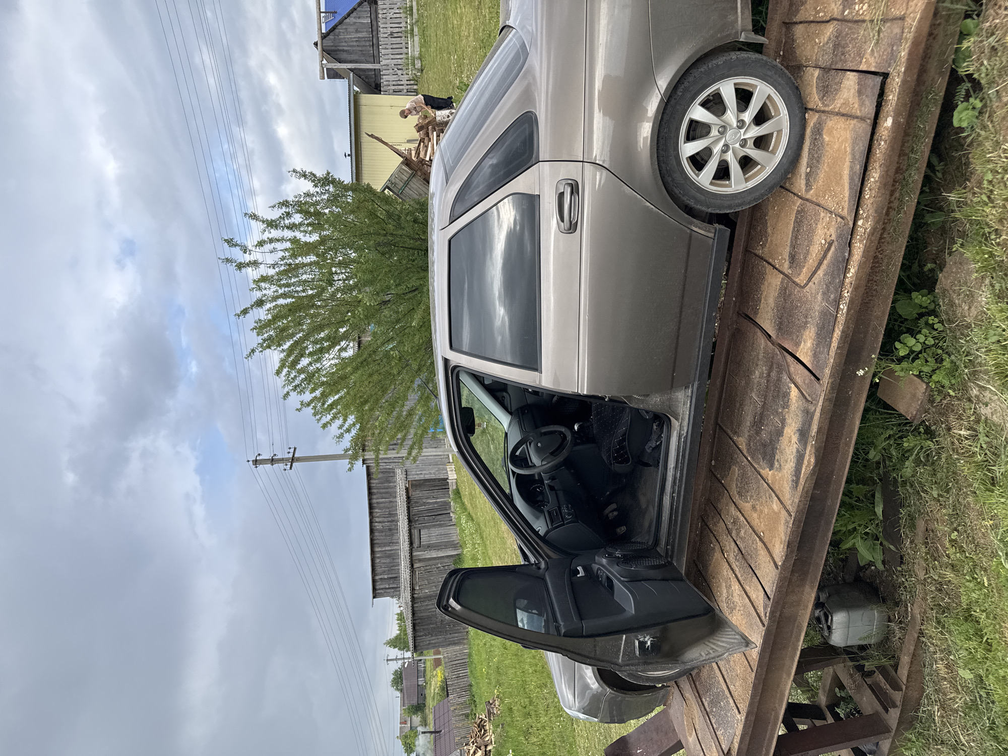 This image captures a rural scene in Бородулино, Russia, under an overcast sky during the daytime. The primary subject is a light brown sedan, possibly a Lada, parked with its front wheels on a rustic, improvised ramp or bridge made of metal and wood, which spans a grassy ditch. The driver's side front and rear doors of the car are wide open, revealing the dark interior, including the steering wheel and floor mats.
In the mid-ground, a person wearing a light shirt and dark pants is visible, standing near a large pile of chopped wood, suggesting they are engaged in outdoor work. The background features several wooden buildings, possibly houses or sheds, and extensive green grass. A tree with vibrant green foliage is partially visible on the left. Overhead, multiple power lines stretch across the cloudy sky, supported by wooden utility poles, indicative of a rural settlement.
A notable detail near the car's front wheel is the textured surface of the ramp, which has a distinct chevron-like pattern. Below the ramp, resting on the grass, is a gray jerry can. The sky is mostly covered by heavy clouds, though some patches of lighter blue are visible, suggesting an overcast day rather than bright sunshine. The scene portrays a glimpse of daily life in a countryside setting, characterized by practical elements like the car on the ramp and the person working with wood. No discernible text is visible anywhere in the image.