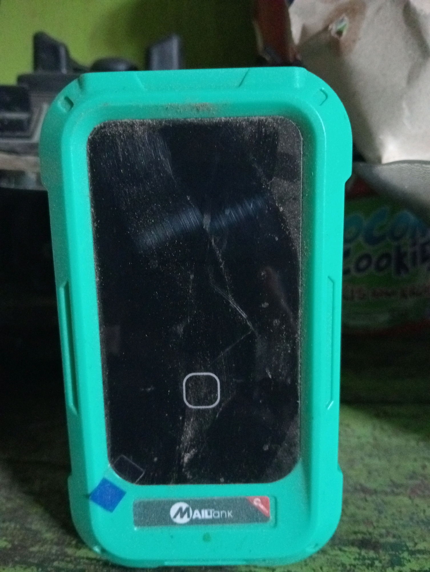 A teal-colored electronic device, likely a point-of-sale terminal, sits on a green, textured surface. The device has a black screen with visible scratches and dust, and a white square icon is visible on the screen. A blue diamond-shaped sticker is affixed to the lower left corner of the device's casing. A logo with the text "MailBank" is present on a silver sticker at the bottom of the device. In the background, out-of-focus objects, including what appears to be a beige bag and dark-colored electronic components, are visible. The environment suggests an indoor setting, possibly a shop or storage area, given the presence of a bagged item and other electronic equipment. The lighting is dim, creating shadows and highlighting the dust on the device. The image was taken in Pagerungan Kecil, Indonesia.