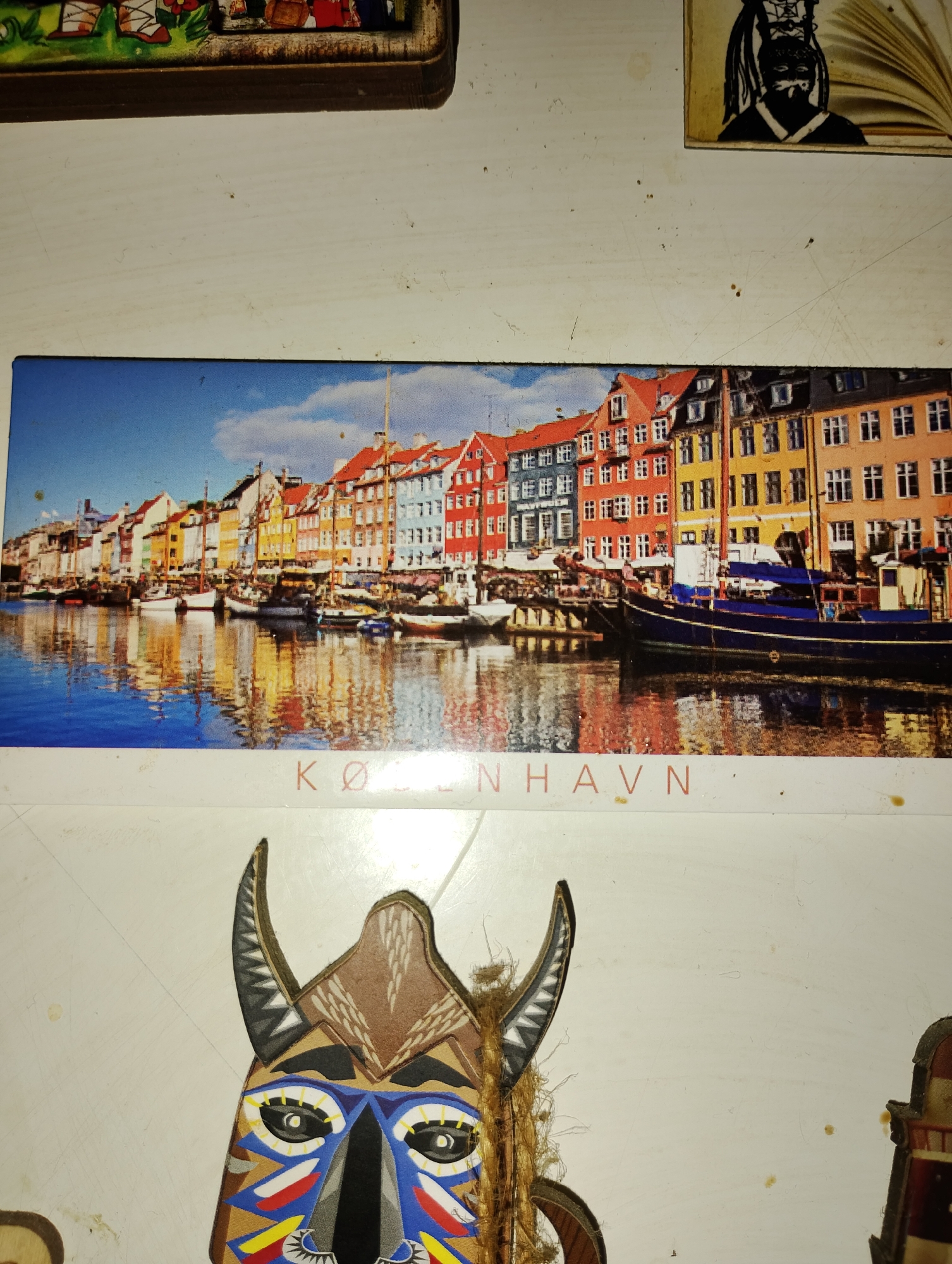 The image displays a collection of items on a white, textured surface. In the upper portion, a small framed image shows a stylized representation of people. To its right, another small framed image depicts a black and white silhouette of a face.
The central and largest element is a rectangular image of Nyhavn, Copenhagen, showing colorful buildings lining a canal with several boats docked. The reflection of the buildings and sky is visible in the water. Below this image, the text "KØ BENHAVN" is printed in red.
The bottom portion of the image features a part of a cutout mask with a tribal design. It has horns, intricate facial markings in blue, yellow, red, and white, and some frayed rope elements. To the far right, a sliver of another item, possibly part of a decorative piece, is visible. The surface appears to be a table or countertop with visible smudges and dust.