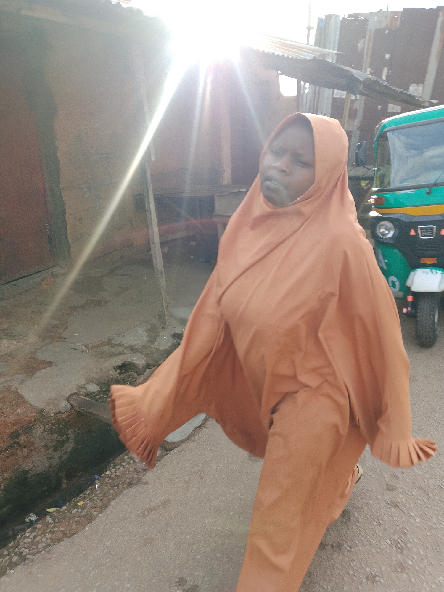 An outdoor, eye-level, medium shot depicts a young Black person walking on a paved road in an urban environment. The individual is wearing a long, orange hijab that covers their entire body. Their face is visible, looking to the right, with a neutral expression. The person's right hand is extended slightly to the side, and the fabric of the hijab is draped over it. The background reveals a rough, unpaved area to the left with a dirt drainage channel filled with debris. Beyond this, a weathered building with a dark brown door stands. To the right of the person, a green and black auto-rickshaw is parked on the road. The scene is illuminated by bright sunlight, casting strong lens flares and highlighting the corrugated metal roof of a structure behind the auto-rickshaw, suggesting a location within Jos, Nigeria, possibly in a less developed area with informal housing and transportation.