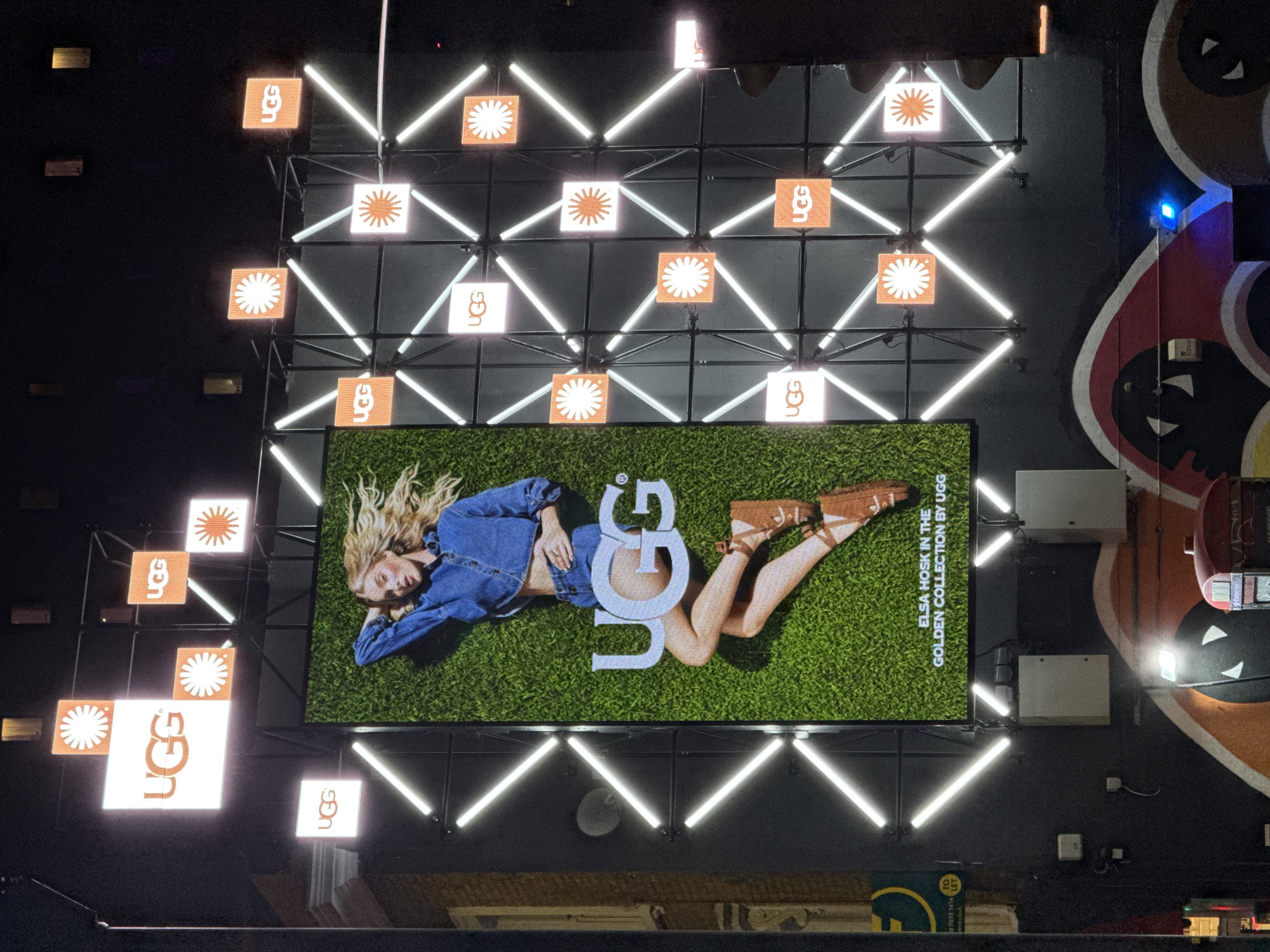 The image captures a large, illuminated UGG advertisement displayed on a building exterior, likely in London, United Kingdom. The sign features a model, Elsa Hosk, lying on a green background, with the UGG logo prominently displayed. The advertisement is part of the "Golden Collection by UGG."
The UGG logo is also featured in several smaller, square panels with flower-like patterns interspersed, creating a grid-like design surrounding the main advertisement. Bright white diagonal light bars intersect the panels, adding to the overall illumination and geometric design.
Adjacent to the UGG display is a dark wall with a large, stylized mural or painting in shades of red, white, and black. Below the UGG display, there's a partial view of a building, suggesting the sign is elevated. A small "To Let" sign is also visible.