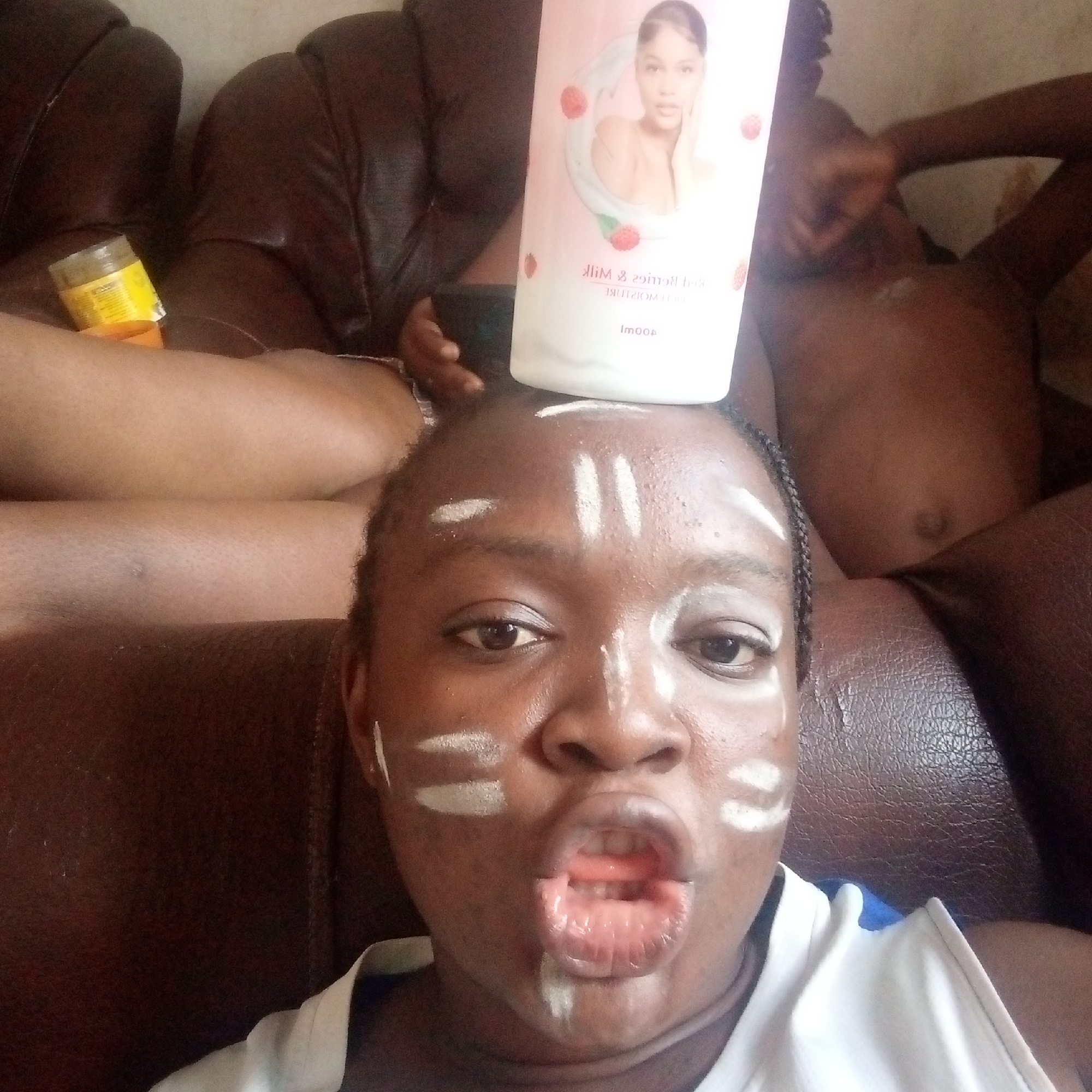 The image depicts a dark-skinned person with white markings on their forehead and cheeks, suggesting face paint or a similar substance. A white container with a product label (featuring a female image and text) is balanced on their head. The person is making a comical face, with their mouth open.
Behind them is a brown leather-like surface, likely a couch. To the left, a yellow container is visible. Another person, whose upper body is visible, is lying on the couch. The setting appears to be indoors, likely in a home. The location in the city of Ogidi, Nigeria, cannot be confirmed.