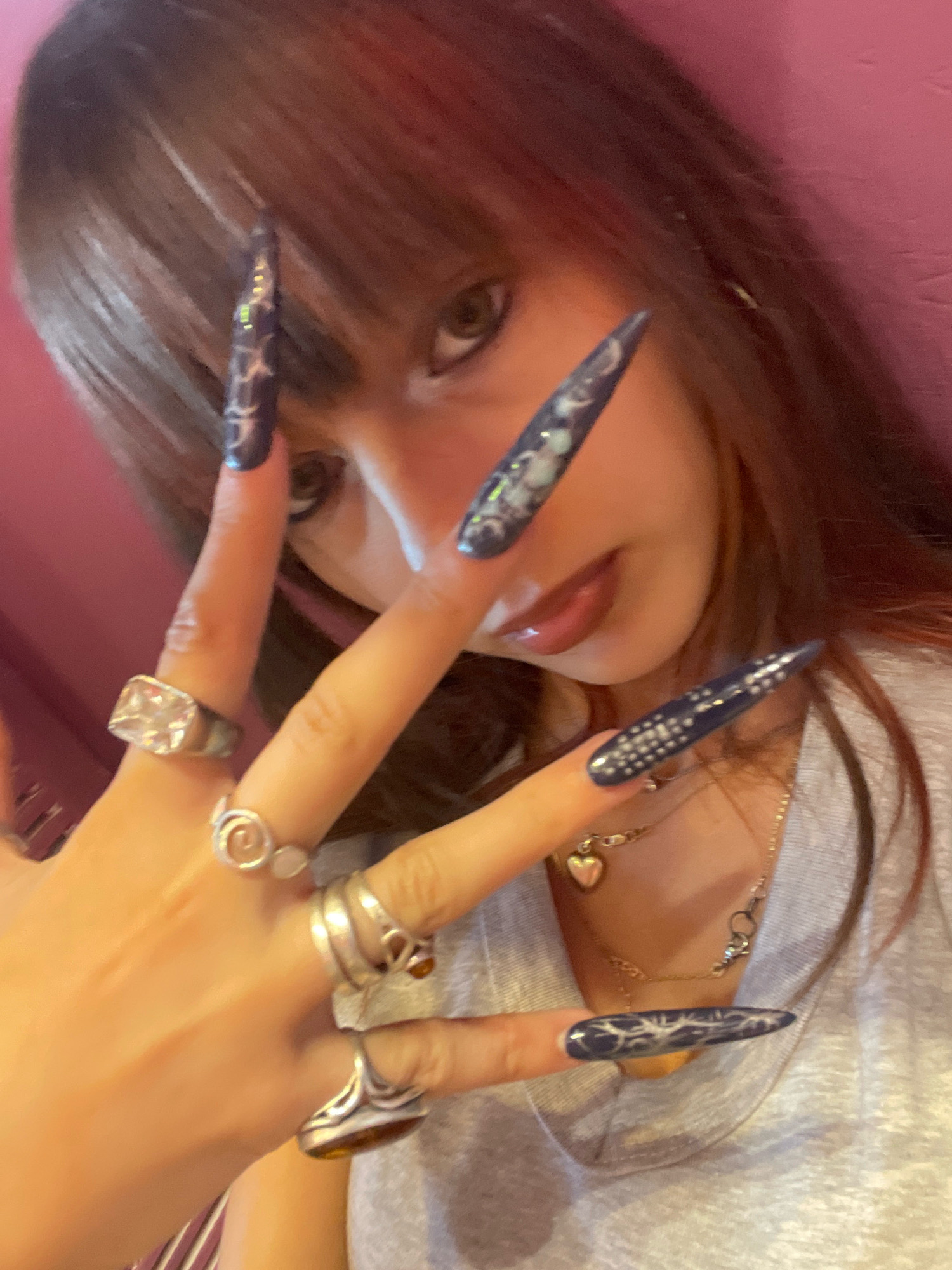 The image shows a close-up of a young woman's face and hands, with a focus on her long, pointed fingernails, which are painted dark blue with intricate white patterns. She is wearing several rings on her fingers, including a large clear stone ring on her index finger and a silver ring with a spiral design on her middle finger. She is also wearing a necklace with a heart-shaped pendant. The background is a blurry pink wall. The scene appears to be indoors, possibly in a home or a studio. There is no information in the image to suggest a specific location in the city of Raanana, Israel.