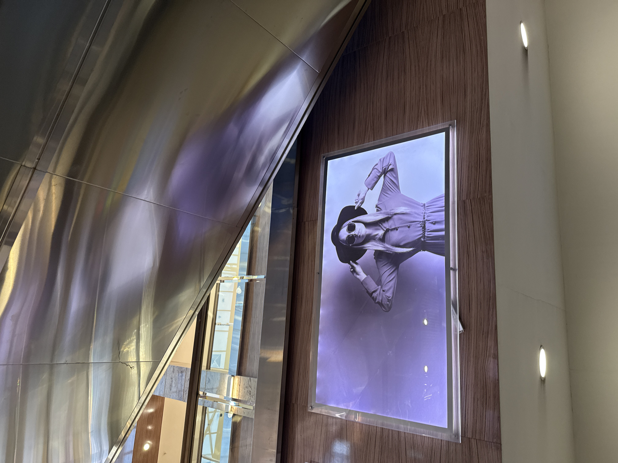 The image shows a vertical shot of a wall with a digital advertisement featuring a woman wearing a wide-brimmed hat and sunglasses, with her hands touching the sides of her hat. The advertisement is illuminated and mounted on a wood-paneled wall. Above the advertisement, a large, curved, metallic structure is visible, reflecting the surroundings. In the upper part of the image, the structure curves downwards, creating a dynamic and modern aesthetic. The metallic structure appears to be part of an escalator or architectural element. The overall environment suggests an indoor public space, possibly a shopping mall or a modern building in Moscow, Russia.