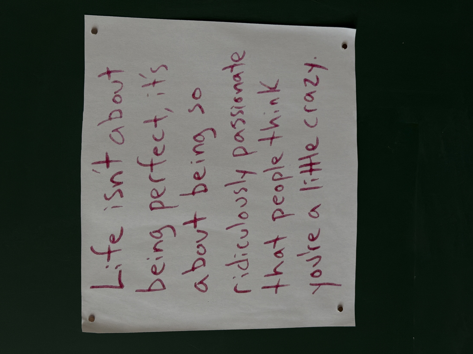 The image shows a piece of white paper with a quote written in dark pink ink. The quote reads, "Life isn't about being perfect, it's about being so ridiculously passionate that people think you're a little crazy." The paper is rectangular and has four small holes, one in each corner. It is placed on a dark green surface. There are no other objects or people visible in the image. The setting is unknown.