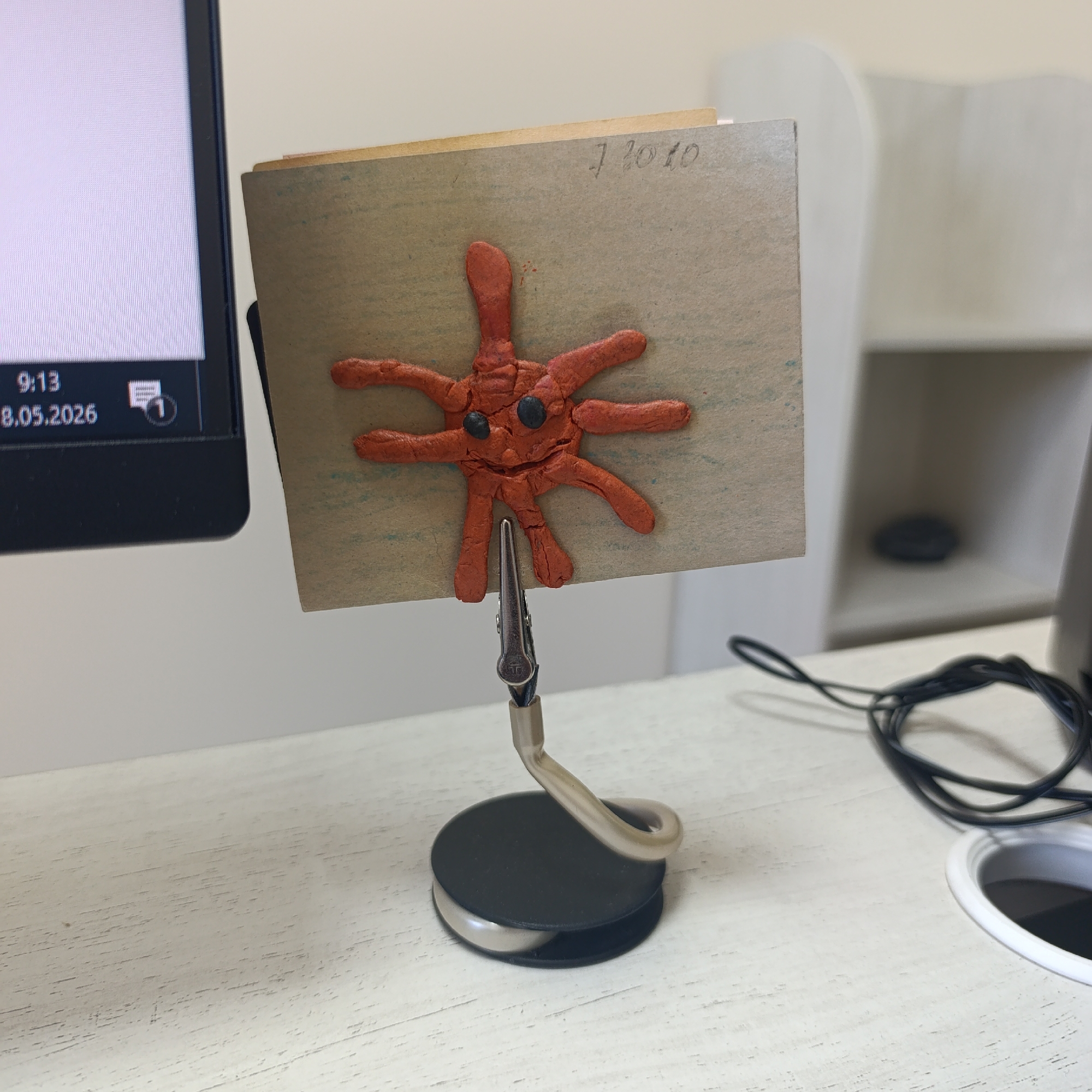 A close-up shows a desk with a computer monitor to the left. An art project on cardboard, featuring an orange clay figure resembling a virus or sea creature with black eyes, is held upright by a clip attached to a base. The cardboard has handwritten numbers, possibly a date, "1 2010". A black cable is visible near a white container on the right. The computer monitor displays the time "9:13" and the date "8.05.2026". No people are visible. There is no information available that this photo was taken in the city of Марковский, Russia.