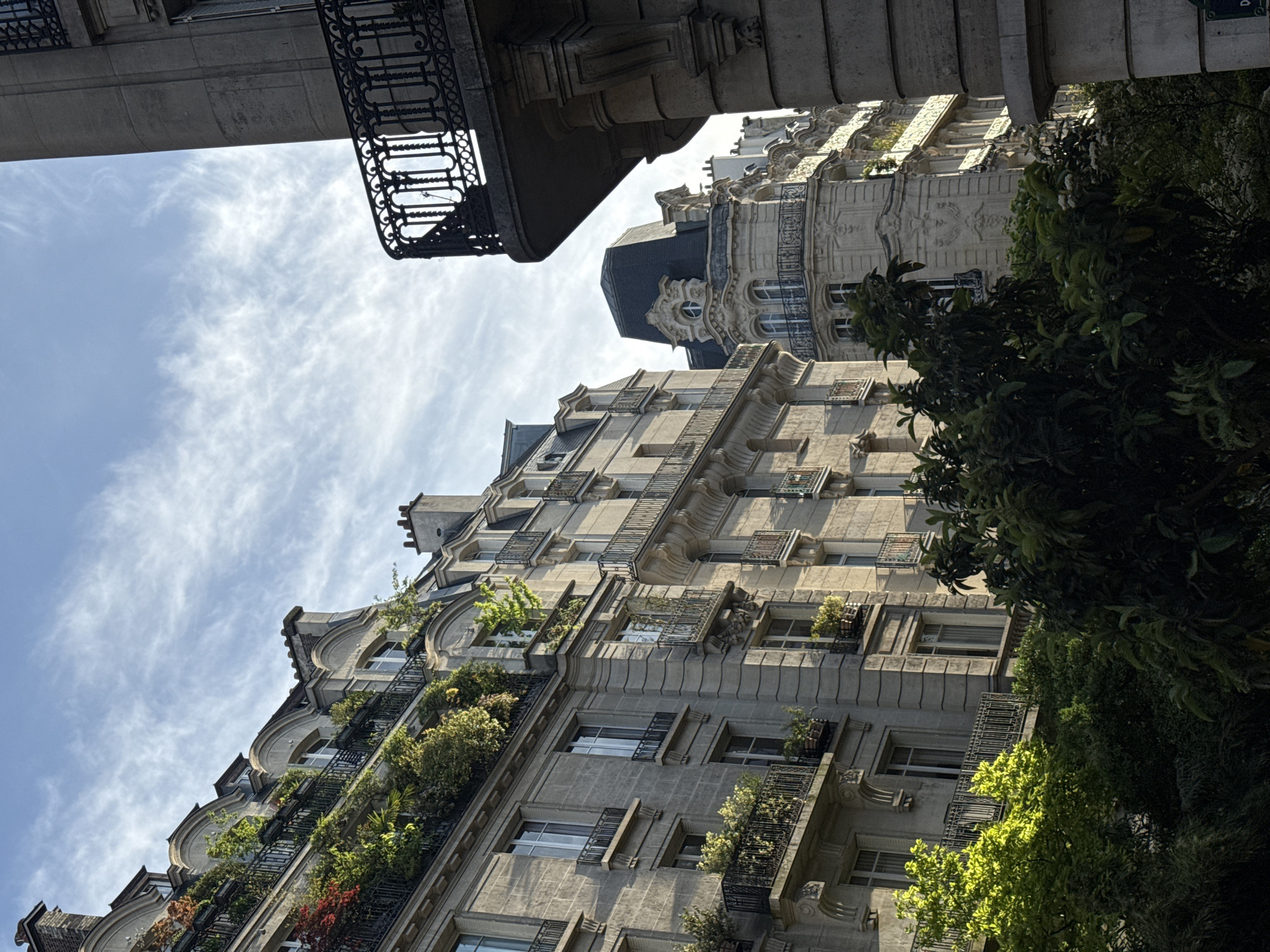 The image presents an upward-looking view of ornate Parisian apartment buildings under a bright, partly cloudy sky. The central focus is a grand, multi-story structure built from light-colored stone or concrete, showcasing characteristic Haussmannian architectural style. Its facade features numerous balconies with intricate black wrought iron railings, many of which are adorned with a variety of lush green and some reddish plants and shrubs, creating a verdant effect across the building. Decorative carvings, cornices, and elegant window frames add to the building's intricate detailing.
To the right, dense green trees partially obscure parts of the buildings, creating natural shadows and highlights where sunlight filters through the leaves. Above and behind the main building, another older, highly decorative building with a dark mansard roof and ornate windows is visible, contributing to the dense urban fabric. From the top left, a heavy, dark metal balcony and the underside of an upper building structure intrude into the frame, emphasizing the cramped, towering perspective.
The sky is a clear blue with scattered wispy white cirrus clouds, indicating fair weather. Strong sunlight illuminates the buildings, casting distinct shadows and highlighting architectural features, suggesting a time of day like morning or late afternoon. No people are visible. A small, partially obscured blue street sign is barely discernible on one of the buildings at the far top right. The scene evokes a sense of historical grandeur and urban beauty, typical of Paris, France, where architecture and greenery harmoniously coexist.
