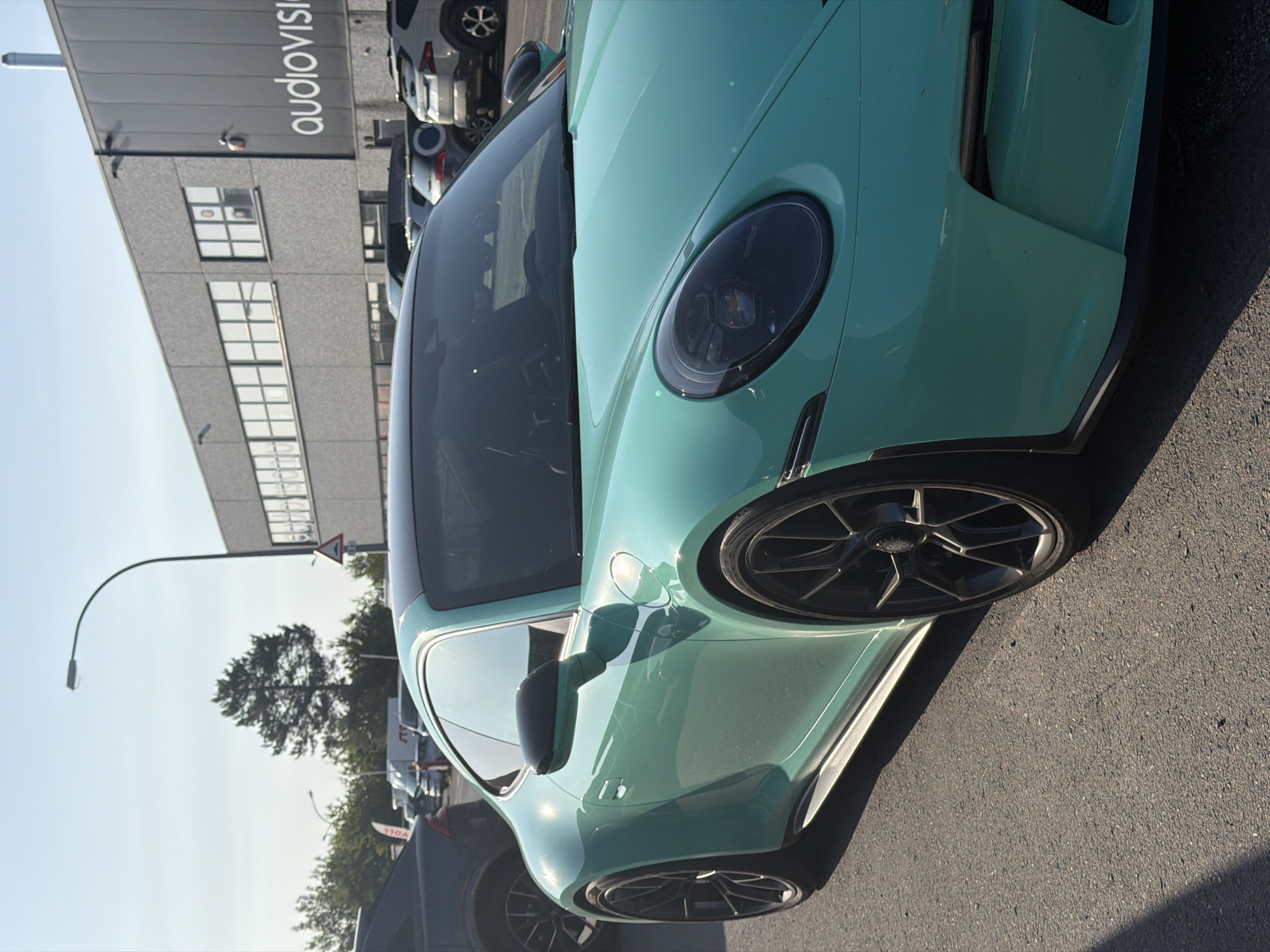 This is a medium-shot, eye-level, outdoor photograph taken on a sunny day. The primary subject is a bright turquoise Porsche 911 GT3 parked on an asphalt surface, likely a street or parking lot. The car is angled from the front left, with its driver's side facing the camera. The car features black wheels and a black roof.
In the background, a modern, grey, industrial-style building is visible, with multiple windows and the large white text "audiovision" on its facade. A streetlamp with a curved arm is positioned behind and to the left of the Porsche, extending towards the top of the frame. Several other vehicles, including a dark-colored SUV to the left and other cars in the distance, are parked near the building. A single tree with dark green foliage stands between the streetlamp and the building. A triangular warning sign is visible on a pole near the building. The sky is clear and light blue.
The location is identified as Leudelange, Luxembourg. There are no people visible in the image. The scene appears to be a commercial or industrial area, possibly a business park or a dealership.