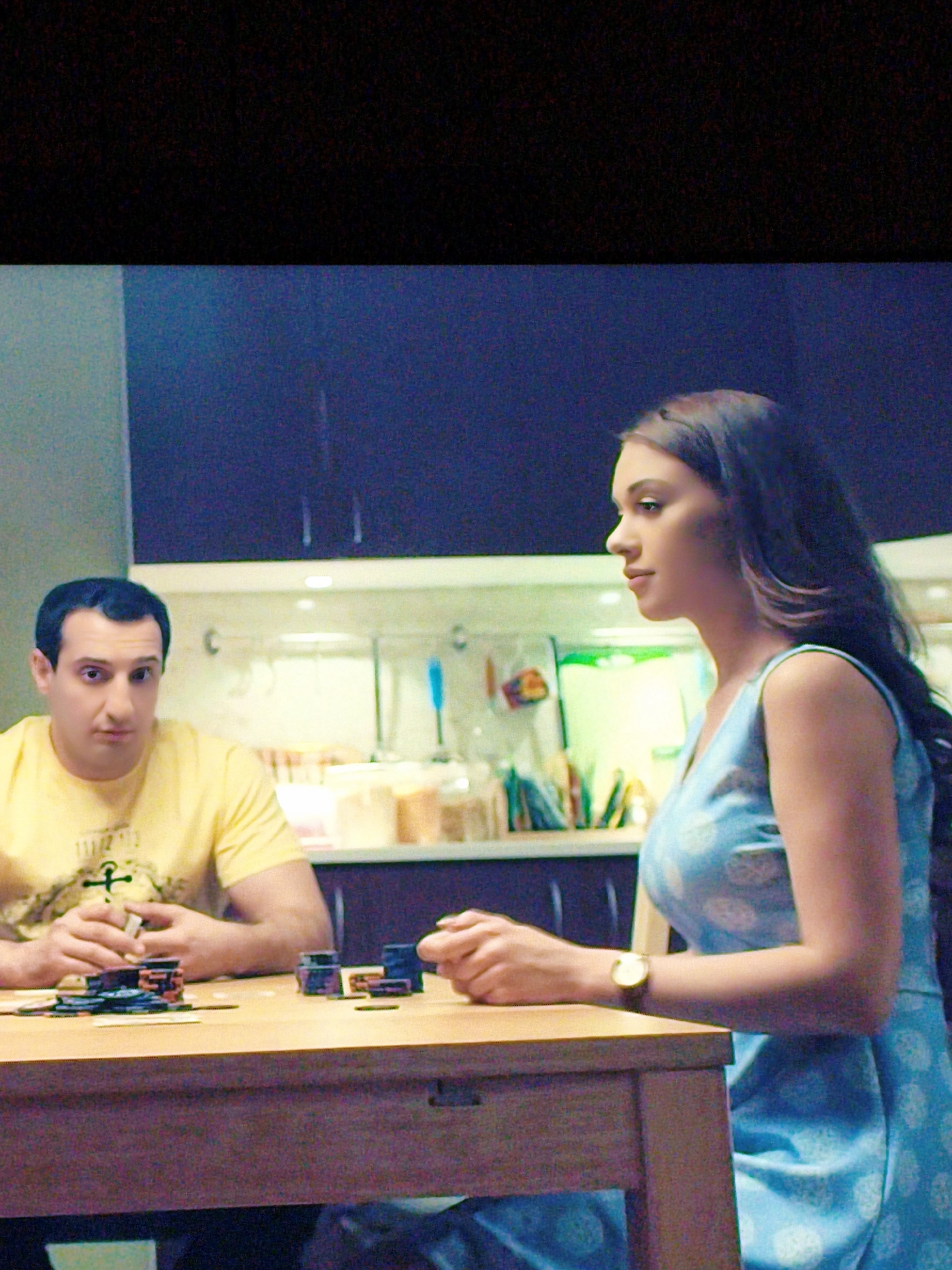 In an indoor setting, possibly a kitchen or dining area, two individuals are seated at a wooden table. The man, wearing a yellow t-shirt, is on the left, his hands resting on the table in front of a stack of poker chips and cards. He appears to be mid-conversation, looking slightly to his left. On the right, a woman in a blue patterned dress is turned away from the camera, facing the man. She is looking to her left, her left arm resting on the table. A gold-toned wristwatch is visible on her left wrist. The background reveals kitchen cabinets and shelves containing various items, including jars and utensils. The overall lighting suggests an indoor environment, with no visible indication of Vladivostok, Russia.