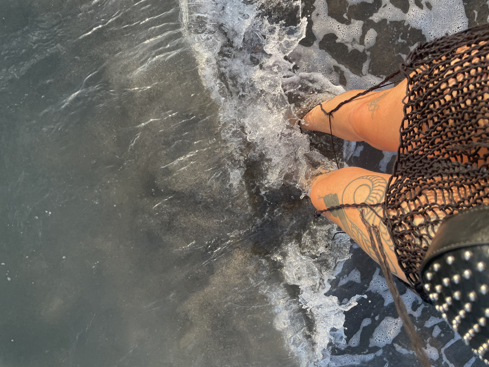 In Donji Štoj, Montenegro, a person stands with their feet in the shallow water of the Adriatic Sea. The water is murky and dark, with waves crashing gently against their legs. The person has tattoos on their legs and is wearing a black mesh skirt over dark shorts and a studded leather top. The sun appears to be low in the sky, casting a warm glow on the scene. The shoreline is not visible, indicating the person is standing in the water with their back to the shore.