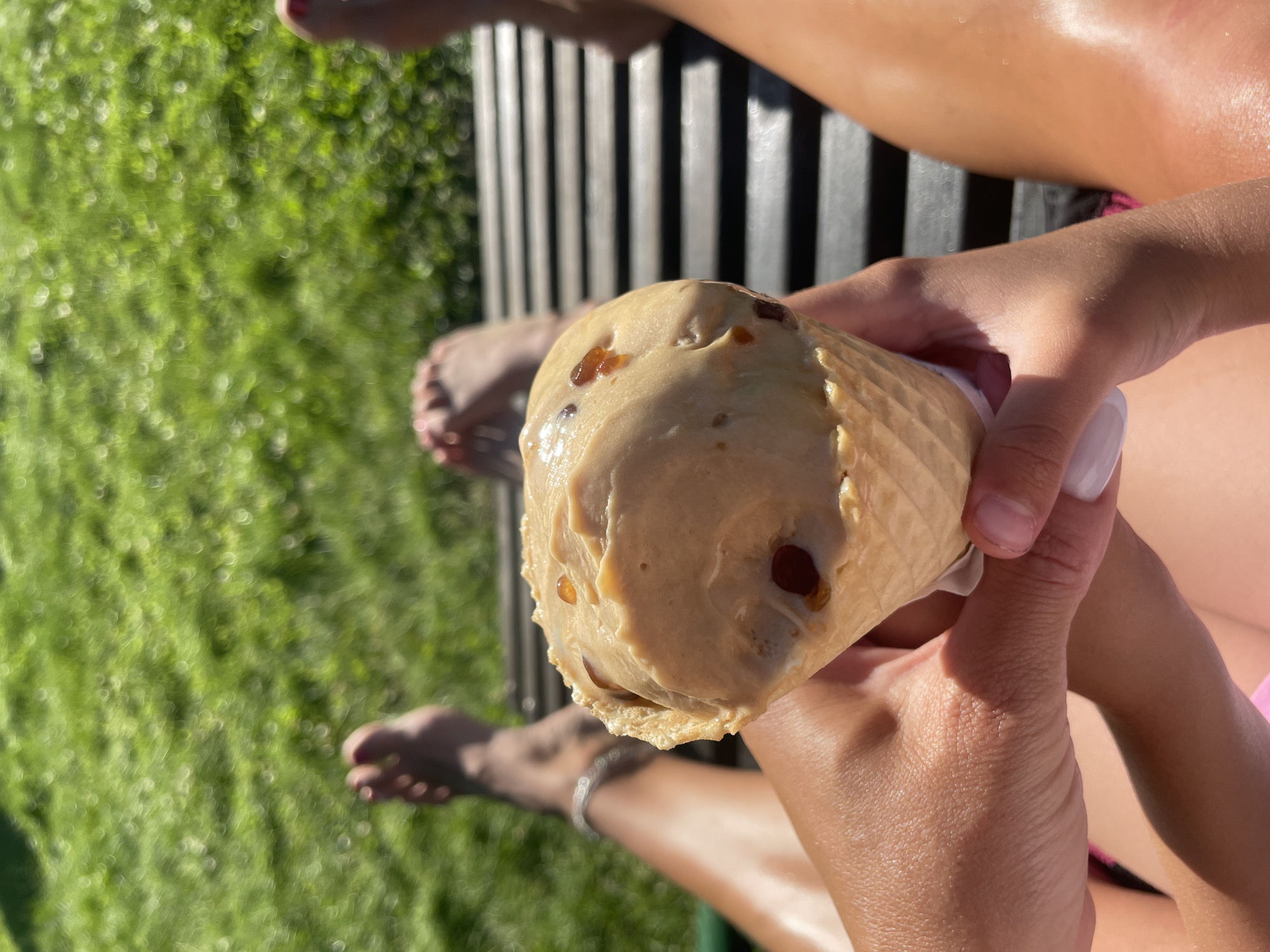 The image shows a close-up of two hands holding an ice cream cone. The ice cream appears to be a light brown color with chocolate chips. The cone is a waffle cone. In the background, out of focus, is a person's legs and feet, and a grassy area. There are also dark vertical lines, possibly part of a bench or railing, in the background. The lighting suggests it is daytime and sunny. The scene appears to be outdoors, possibly in a park or backyard. No text is visible in the image.