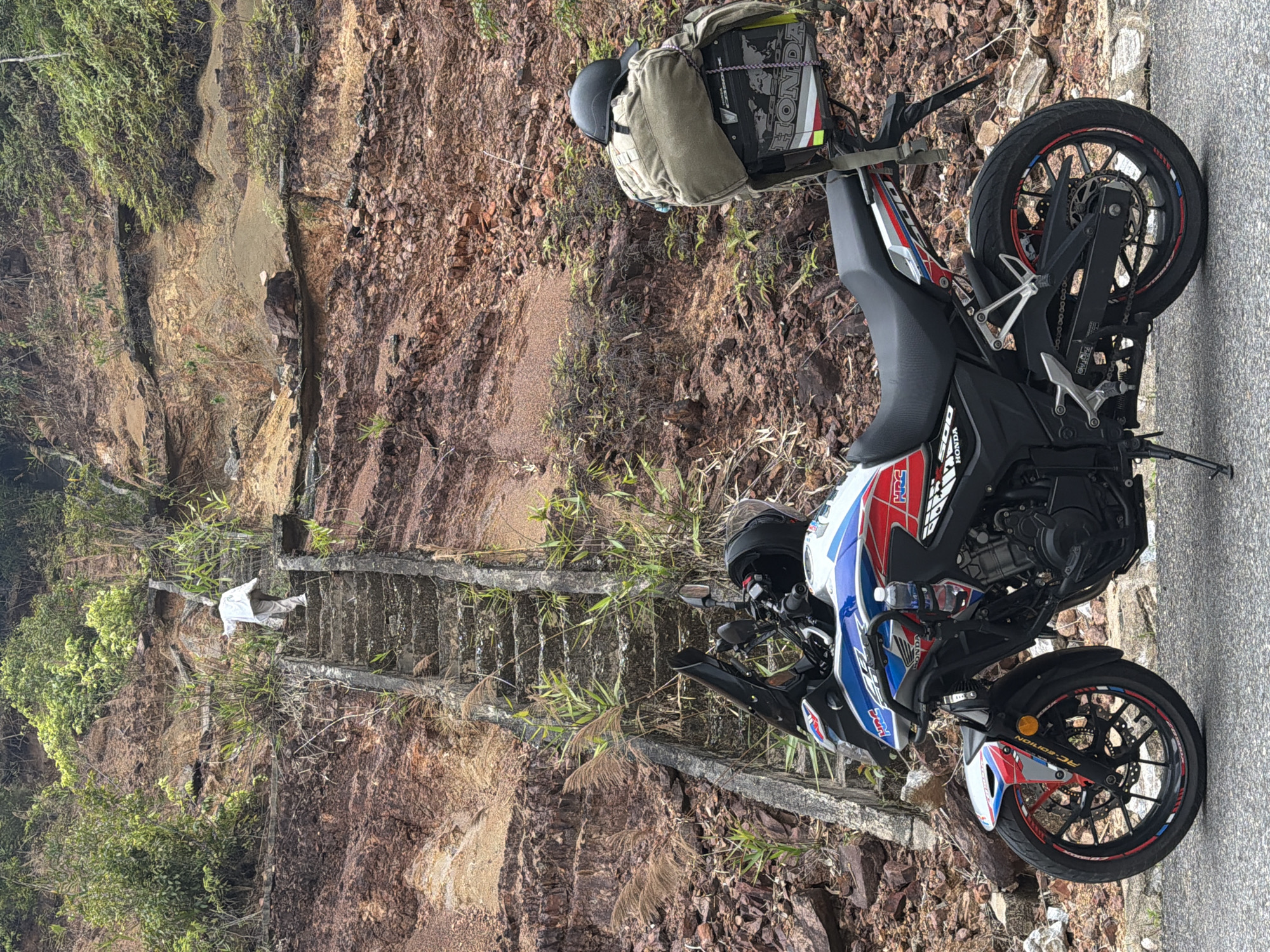 The image depicts a motorcycle parked on the side of a road in a mountainous area. The motorcycle is a Honda CB500X with a red, white, and blue color scheme. A backpack and a helmet are secured to the back of the motorcycle.
To the left of the motorcycle, there is a steep, rocky hillside. A set of concrete stairs with vegetation growing on the sides leads up the hillside. A person wearing white is walking up the stairs.
The image was taken in a natural environment, which could possibly be in the city of Đà Nẵng, Vietnam.