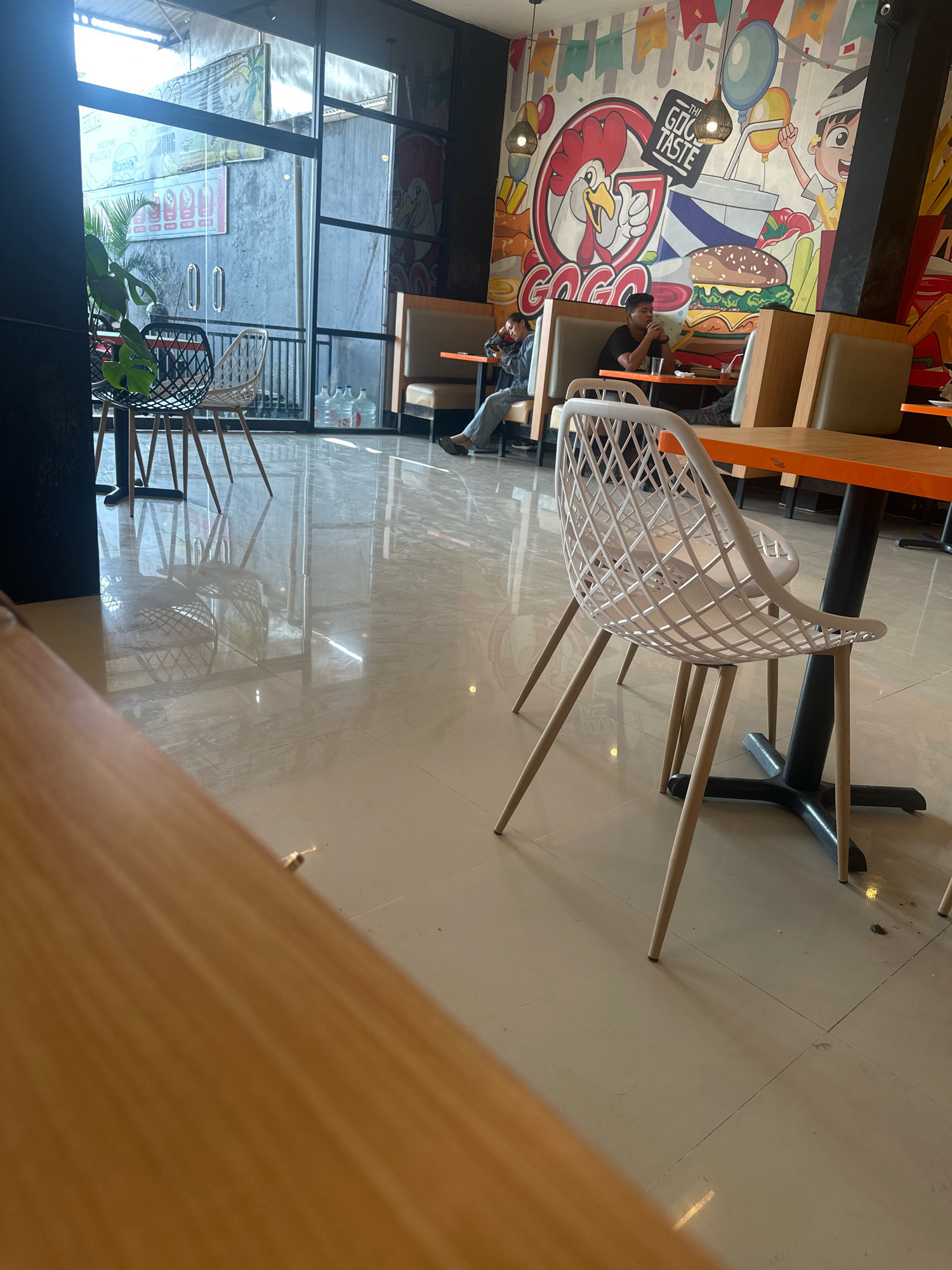 The image depicts the interior of a restaurant in Kerambitan, Indonesia, characterized by a modern and vibrant design. In the foreground, a light-colored wooden table is partially visible, suggesting a seating area. Several white, modern chairs with intricate woven designs are placed around tables. One such chair is prominently featured in the mid-ground, positioned near an orange-topped table with a black pedestal base.
Further into the restaurant, two individuals are seated at tables. One person, wearing a dark shirt, is seated at a booth-style bench, looking down and appearing engaged with their meal. Another individual, in a grey hoodie and jeans, is seated at a similar booth, also looking down.
The background features a large, colorful mural on the wall, depicting cartoonish characters, a fried chicken advertisement, and burgers, creating a lively atmosphere. Large glass windows are visible on the left side of the image, offering a glimpse of the exterior, which includes what appears to be a wall and some greenery. The floor is tiled and highly reflective, mirroring the overhead lighting and furniture. Several large water bottles are visible through the windows. The overall impression is that of a casual dining establishment.