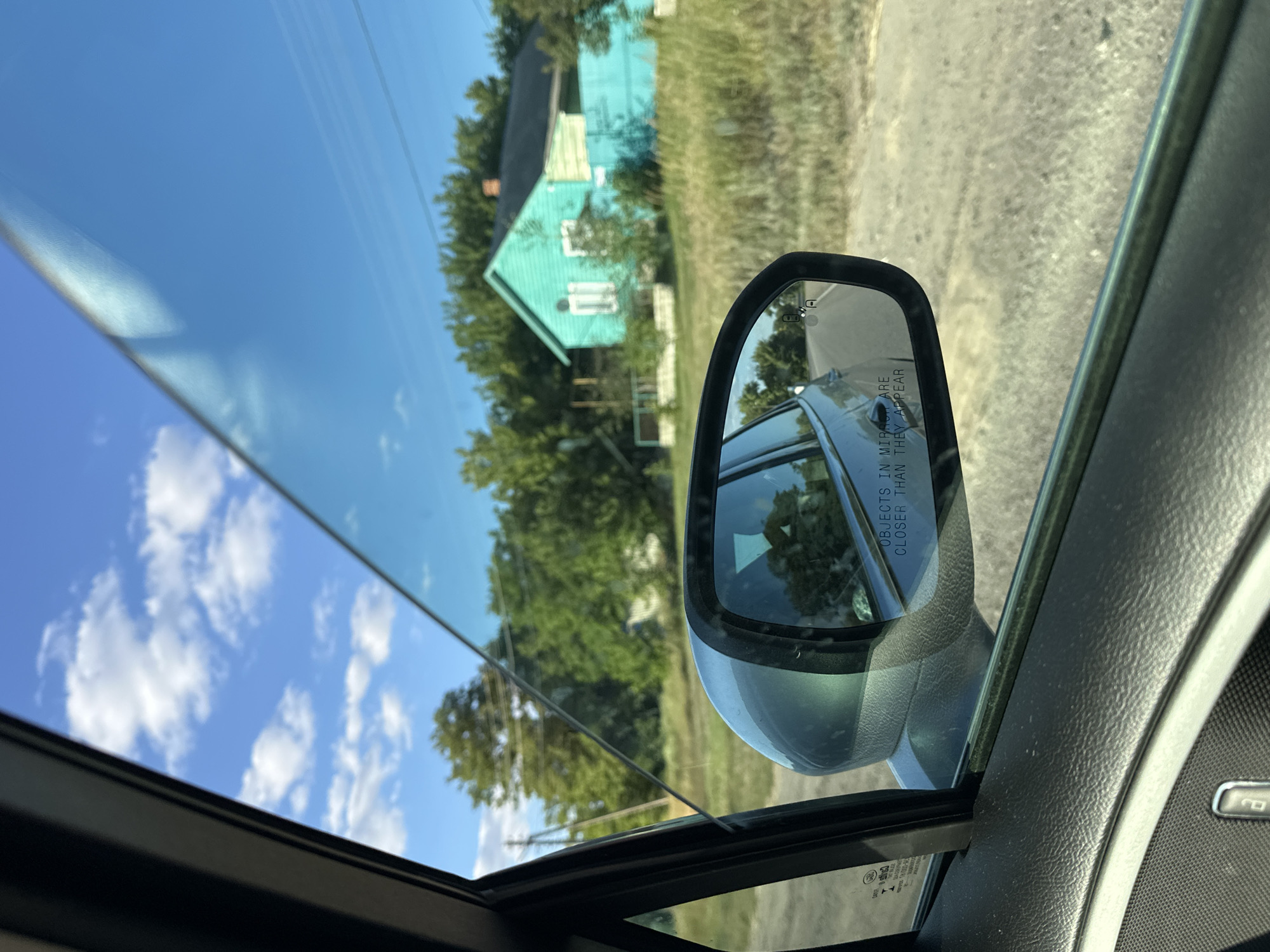 The image captures a view from inside a car, looking out the side window. The scene depicts a rural setting under a clear blue sky with scattered white clouds. A teal-colored house with a black roof and white-framed windows is visible in the background, bordered by a matching teal fence. The house is surrounded by lush green trees and tall, dry grass. A dirt road is visible in the foreground, and a side-view mirror reflects the road behind the car. The interior of the car, including the door panel and window frame, is partially visible. The mirror contains the text "OBJECTS IN MIRROR ARE CLOSER THAN THEY APPEAR." The overall impression is a snapshot of a journey through a quiet, sun-drenched countryside, possibly in the village of Novotolucheyevo, Russia, given the context provided. There are no people or visible actions occurring within the frame.