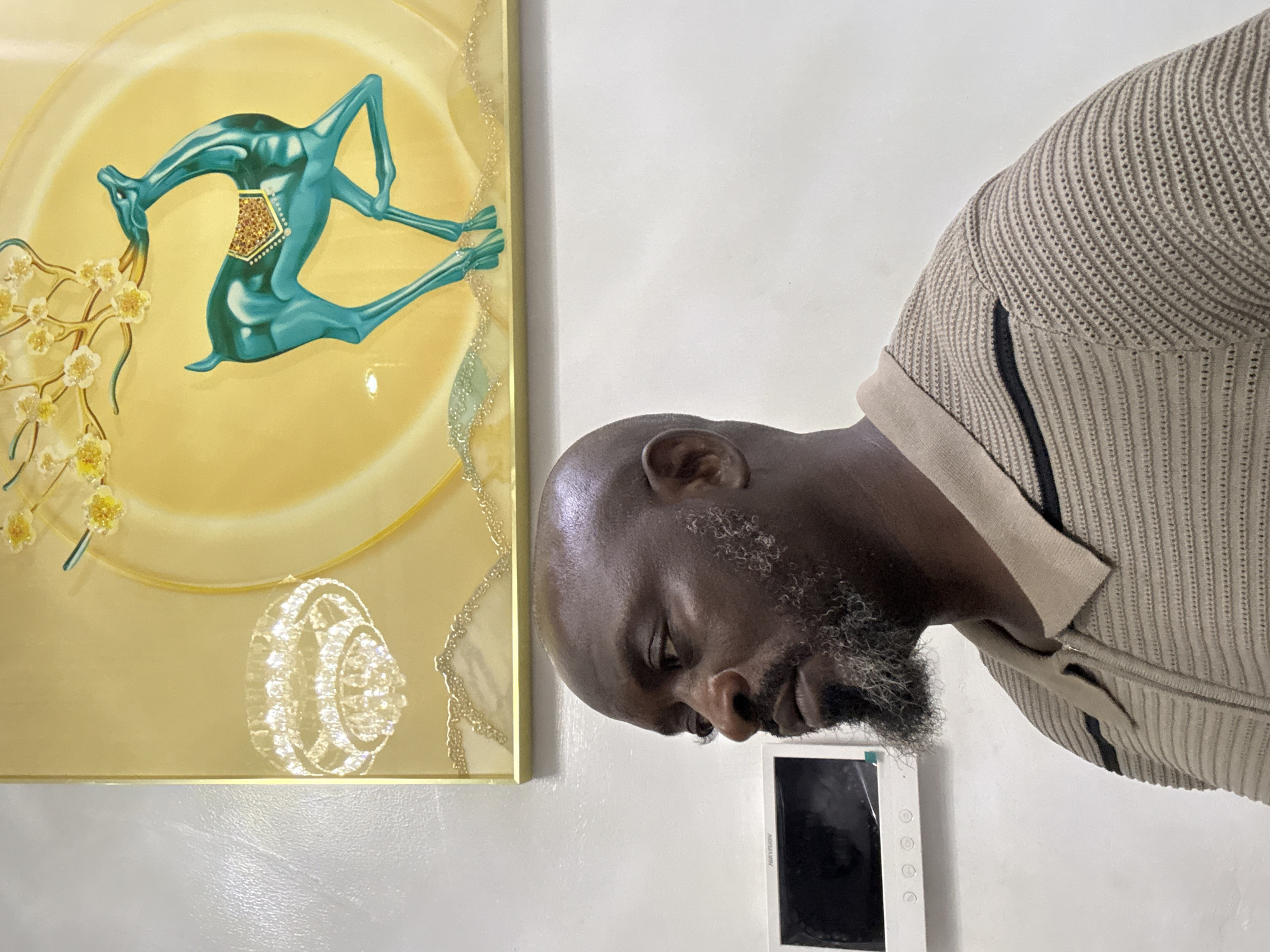 The image captures a close-up shot of a Black man with a bald head and a salt-and-pepper beard. He is wearing a tan knitted polo shirt with black stripes along the sides. He is positioned near a wall, with an artwork hanging on it to his left. The artwork appears to be a stylized, decorative piece with a golden background and a teal figure on it. A white device with a screen is mounted below the man's face. The location is likely an interior space, possibly in Lekki, Nigeria, given the user's context of the request, but this is not explicitly confirmed by the imagery. The man appears to be looking downwards.