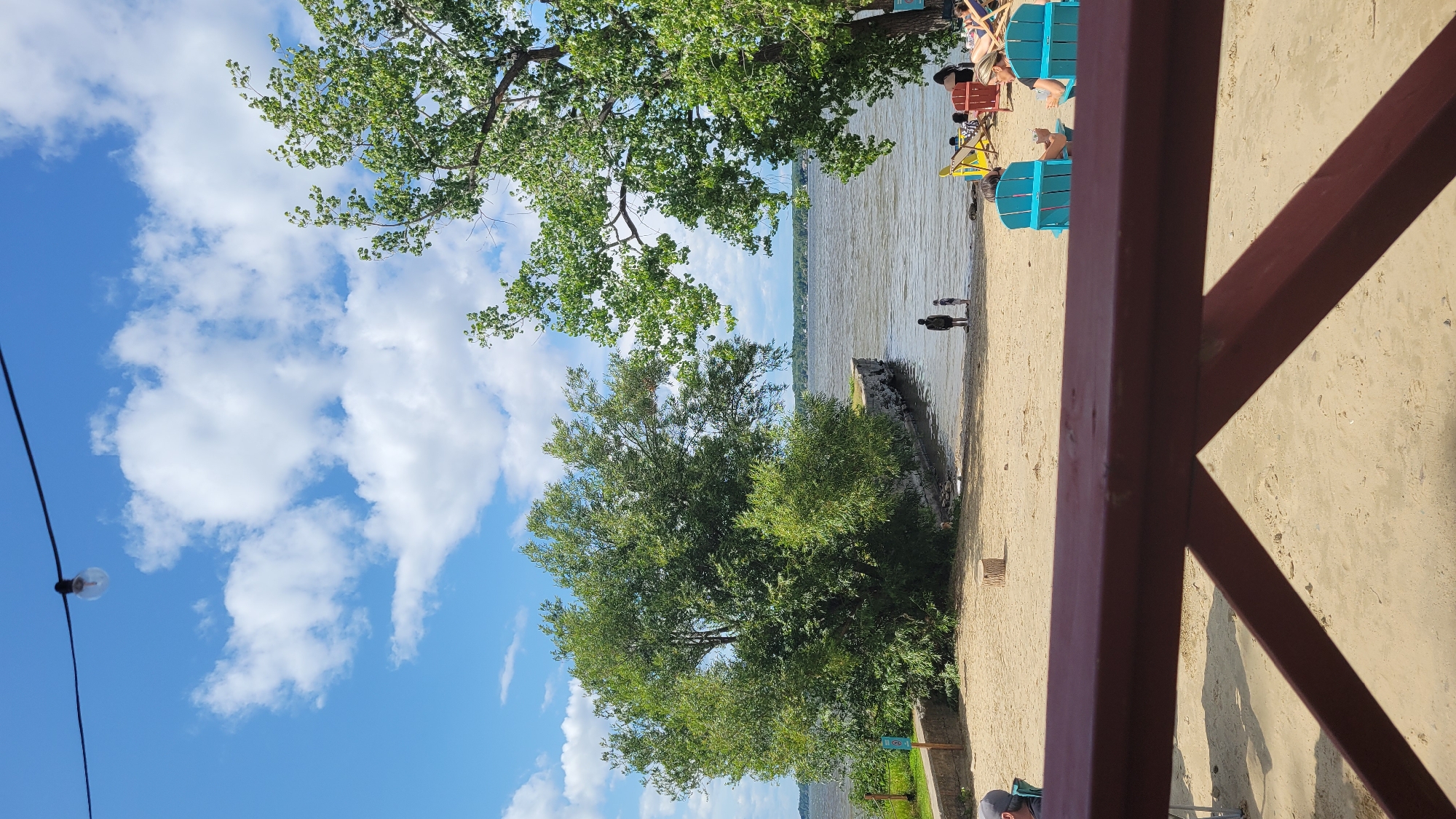 The image depicts a beach scene at the Parc national d'Oka, located in Pointe-des-Cascades, Canada. The foreground is dominated by a maroon-colored railing of a patio or deck, obscuring the lower portion of the sandy beach. Beyond the railing, the beach stretches towards a body of water, likely a lake, with calm, rippling waves.
On the beach, several people are visible. Two individuals, appearing to be an adult and a child, stand near the water's edge, facing the lake. Further back, a group of people are relaxing in beach chairs. Two bright turquoise Adirondack chairs are prominently placed on the sand, each occupied by a person. Other beach chairs, some yellow and some red, are scattered around the area, with more individuals seated in them.
Lush green trees frame the scene, with a large, full tree on the right and another substantial tree on the left, closer to the water. The water extends to the horizon, where a distant treeline is visible under a bright blue sky dotted with fluffy white clouds. A single light bulb hangs from a wire in the upper portion of the frame, suggesting an outdoor lighting fixture. A small, cylindrical tree stump rests on the sand in the middle ground. The overall impression is a relaxed, summer day at a waterfront recreational area.