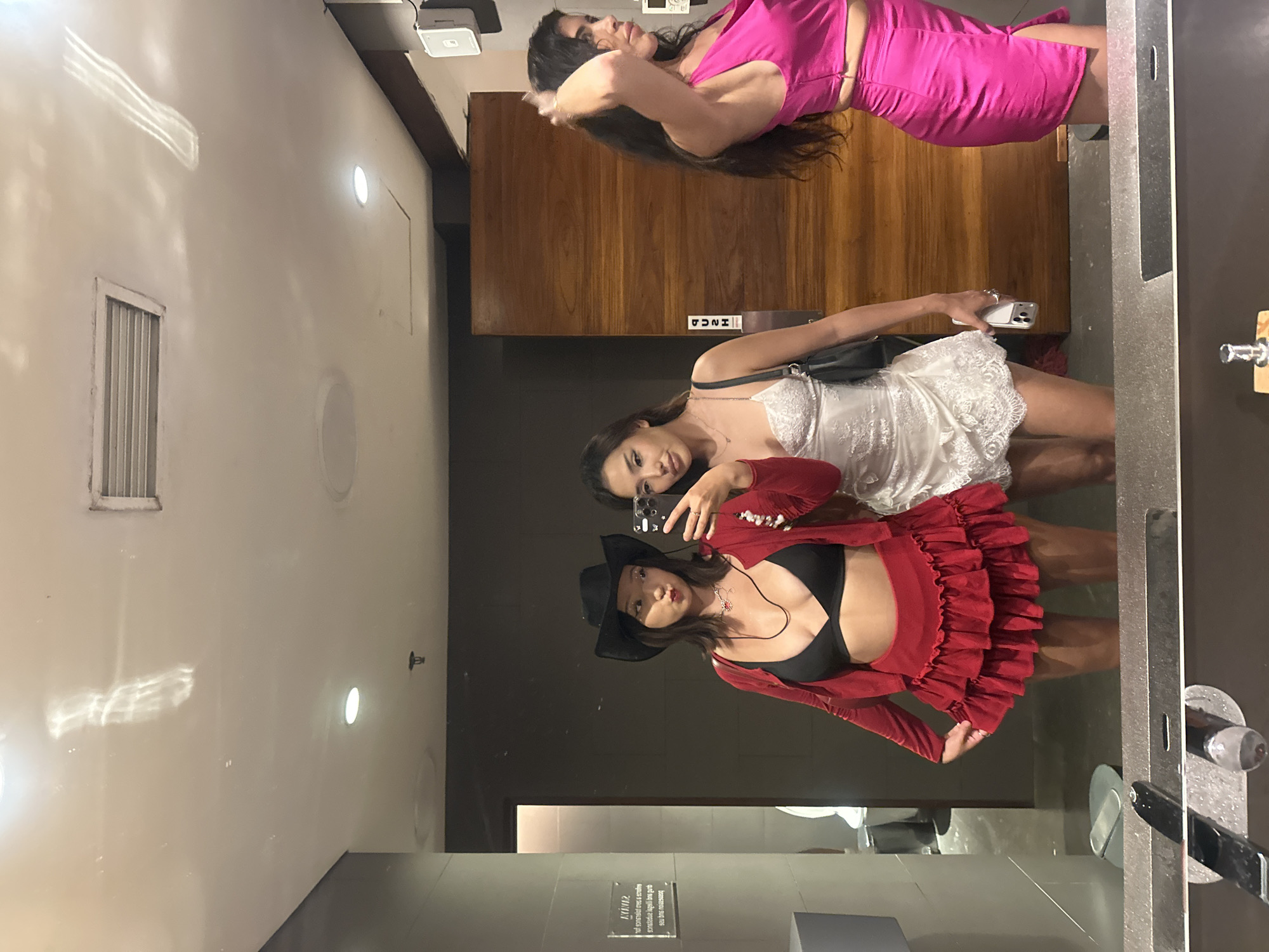 Three women are posing for a mirror selfie in a public restroom. One woman, wearing a pink dress, is lying on a counter or ledge above a wooden panel. Another woman in a white dress is holding a phone, presumably taking the picture. A third woman, wearing a black bra, a red ruffled top and skirt, and a black hat, is posing in front. The restroom has white walls with recessed lighting, a vent, and a wooden door. The location is Kuta Selatan, Indonesia.