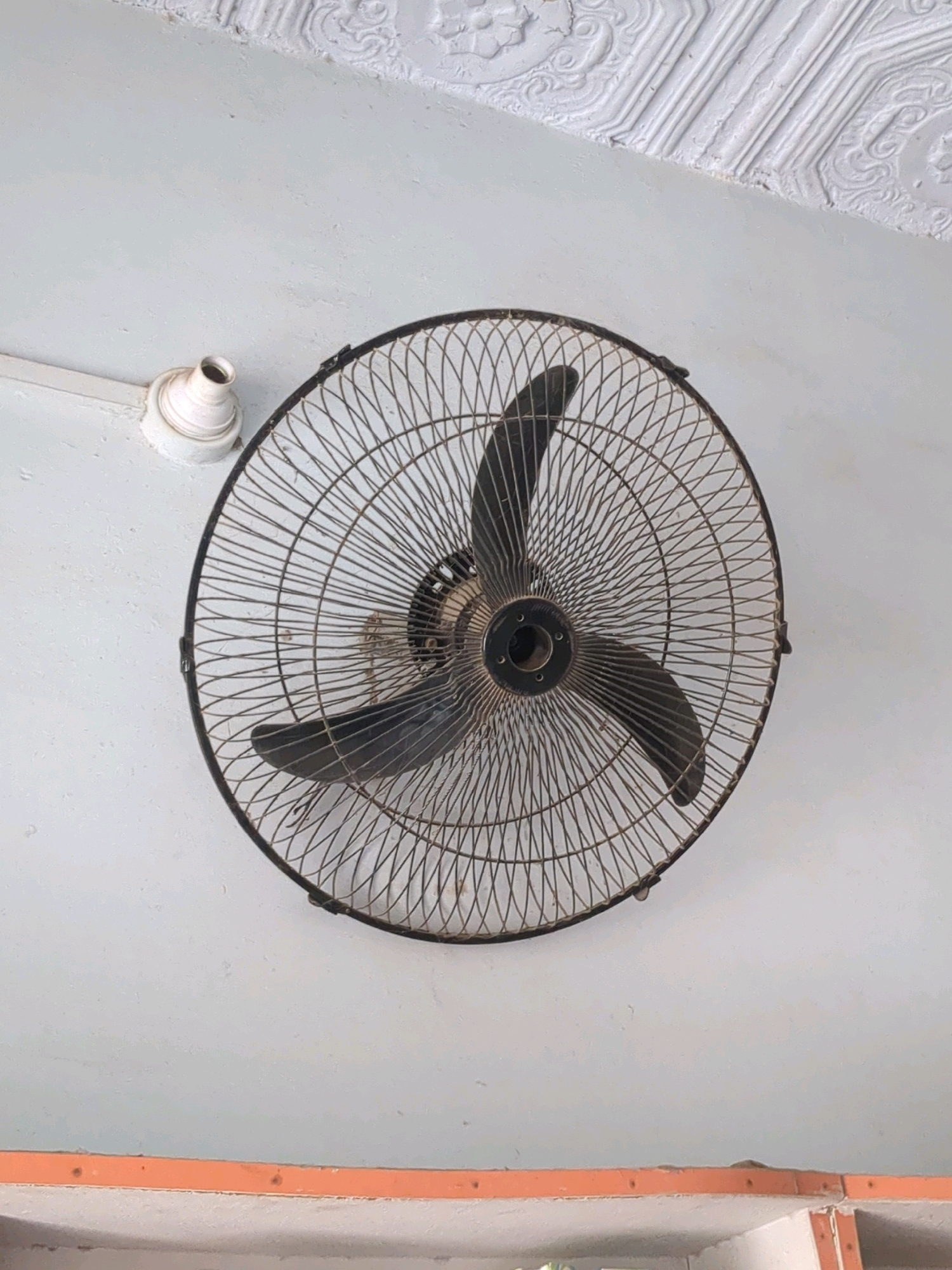 A wall-mounted fan with a metal grill and dark blades is centered in the frame, attached to a light-colored wall. Above the fan, a white ceiling with decorative molding is visible. To the left of the fan, a white electrical conduit is attached to the wall. Below the fan, an orange-colored shelf or ledge runs horizontally across the bottom of the image. The scene appears to be an indoor domestic or commercial setting. There is no visible indication of the specific location within Karachi Division, Pakistan.