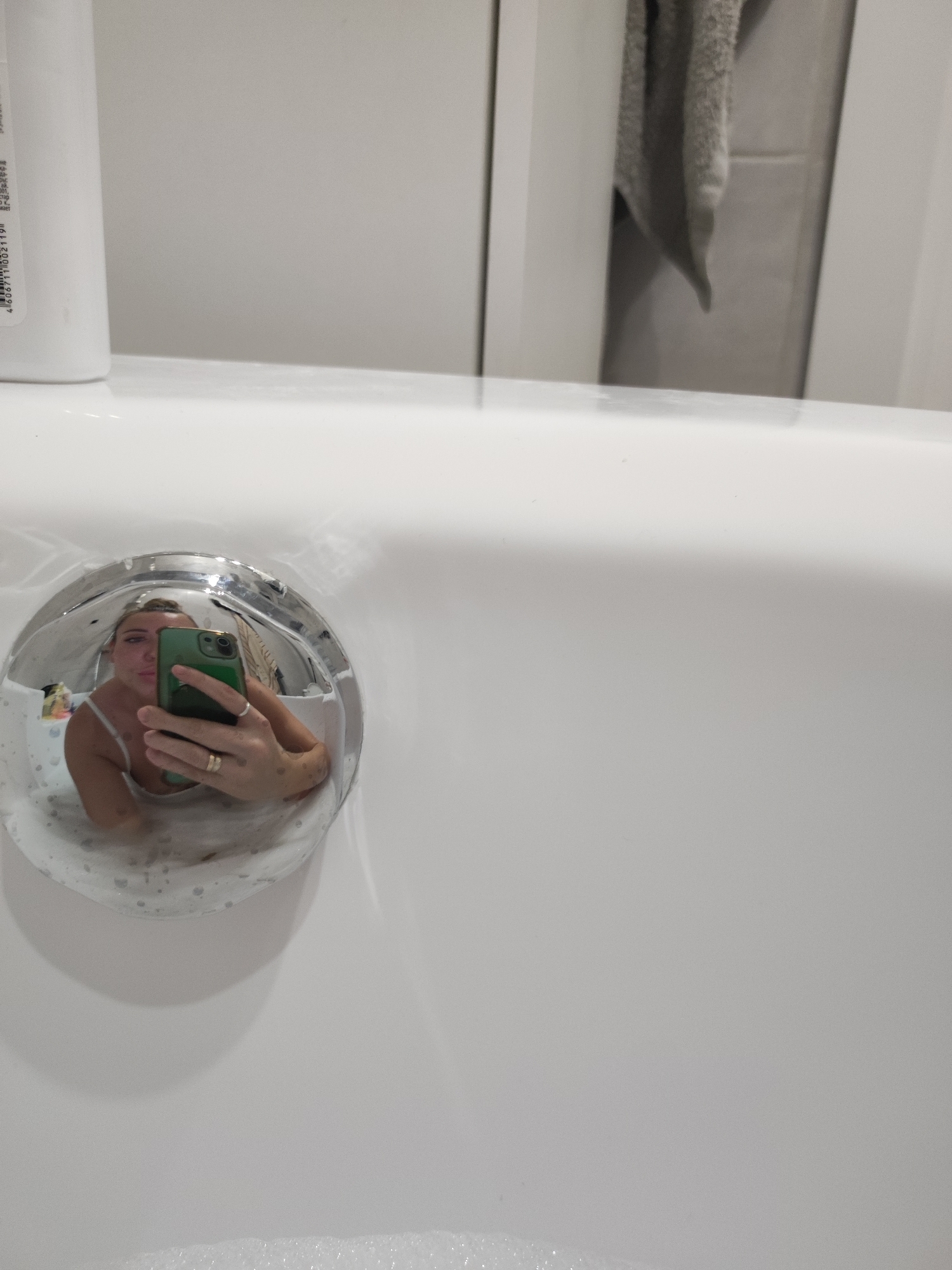 The image is a close-up shot of a white bathtub. The reflection of a person in the bathtub drain is visible. The person is holding a green cell phone. A towel is hanging on the door in the background. A bottle of product is on the left side of the tub. The location is unknown and has no clear indication it is Votkinsk, Russia.