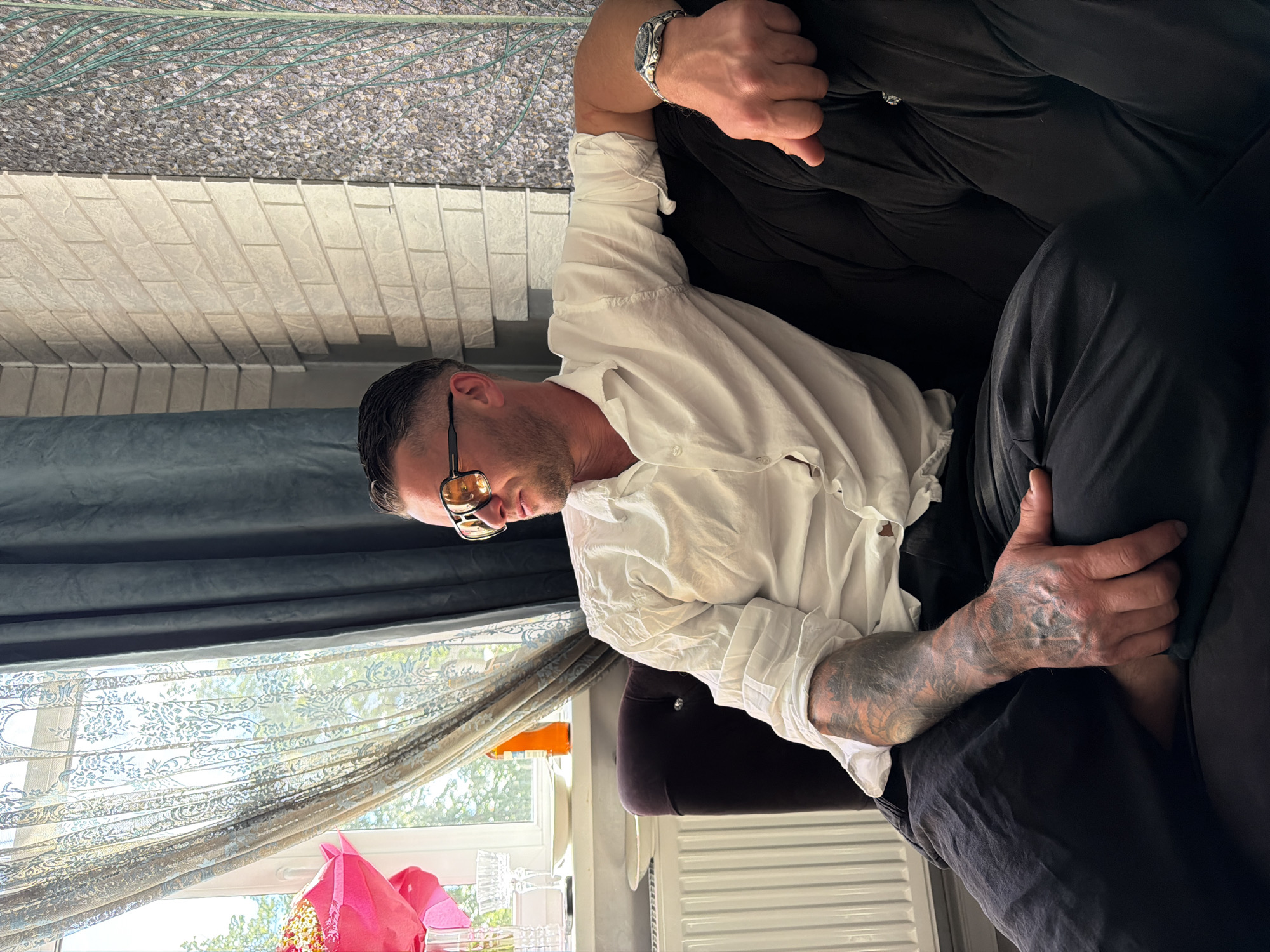 A man with short, dark hair and sunglasses is seated on a dark, tufted sofa. He is wearing a white dress shirt and dark pants. His left arm is resting on the back of the sofa, and his right hand is in his lap. He has extensive tattoos on his left arm and hand. The background shows a window with light blue curtains and a radiator beneath it. To the right of the window, there is a wall with a brick pattern and a textured wallpaper with a floral design. A bright pink balloon bouquet is visible on the windowsill. The scene appears to be indoors, possibly in a residential setting. There is no information in the image to suggest a specific location in the city of Riga, Latvia.