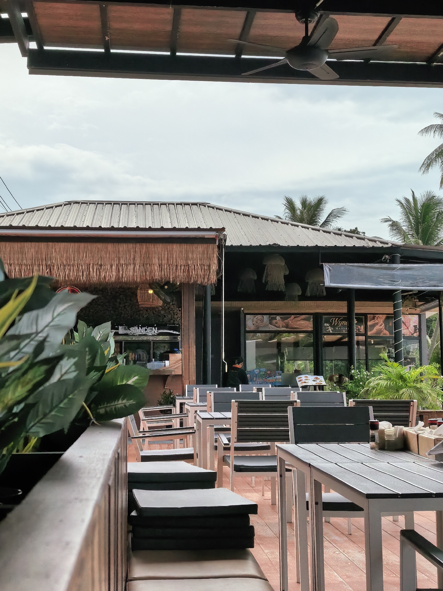 The image depicts an outdoor seating area of a restaurant or massage establishment, likely located in the Ko Samui District of Thailand, given the presence of palm trees and tropical vegetation. The scene is framed by a thatched roof overhang and a ceiling fan, suggesting a warm climate. Rows of tables and chairs are arranged on a wooden deck, some of which are empty, while others are occupied by patrons or staff. In the background, the building features a metal roof and large windows displaying advertisements for massages. A person in dark clothing is seated at one of the tables, facing away from the camera. The overall atmosphere suggests a relaxed, tropical setting.