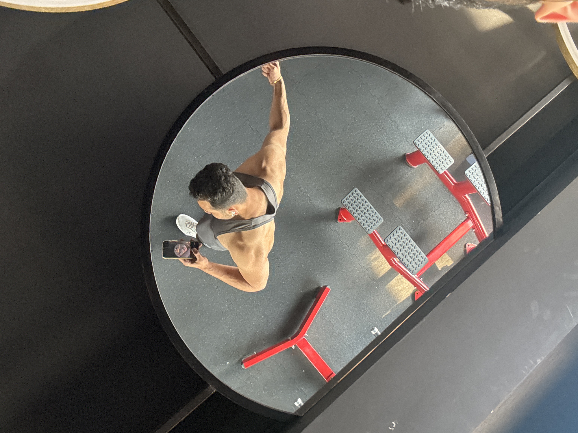 A selfie of a man is reflected in a large, round mirror in a gym setting. The man is muscular, wearing a grey tank top and white sneakers, holding a smartphone. He is standing in front of a red weight-lifting machine, with black flooring visible around him. The location is possibly Kolkata, India.