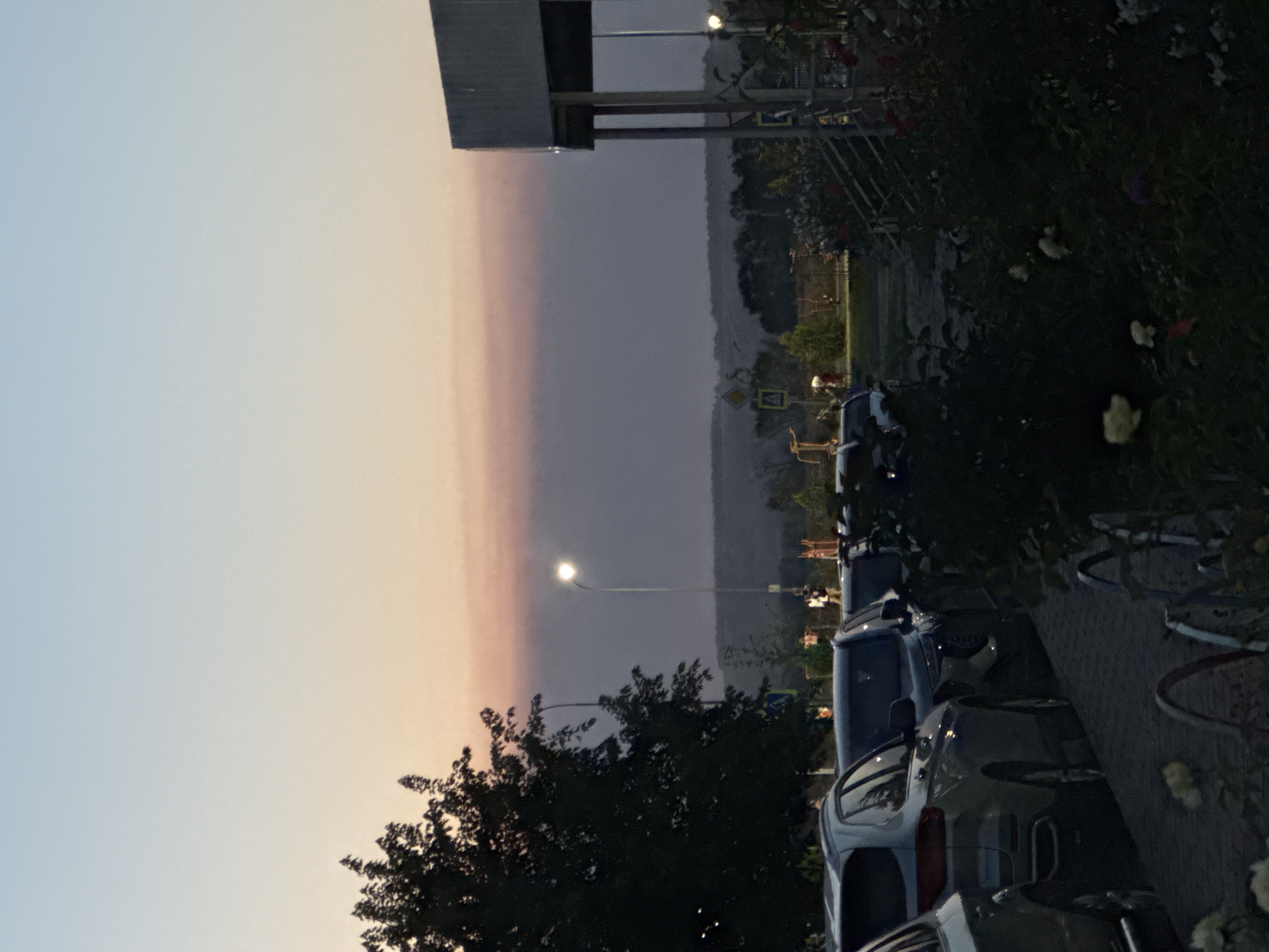 The image shows a parking lot in an urban area at dusk. Several cars are parked in a row on the left side of the image. In the foreground, a tree with dark foliage is visible on the left, and a building with a covered walkway is on the right. A streetlamp with a glowing light is positioned between the tree and the building. The sky is a gradient of pale blue to a soft pinkish-orange haze, indicating the setting sun. In the background, there are rolling hills covered in trees, suggesting a suburban or exurban setting. A few people are walking in the middle ground. The overall lighting is dim, consistent with twilight.