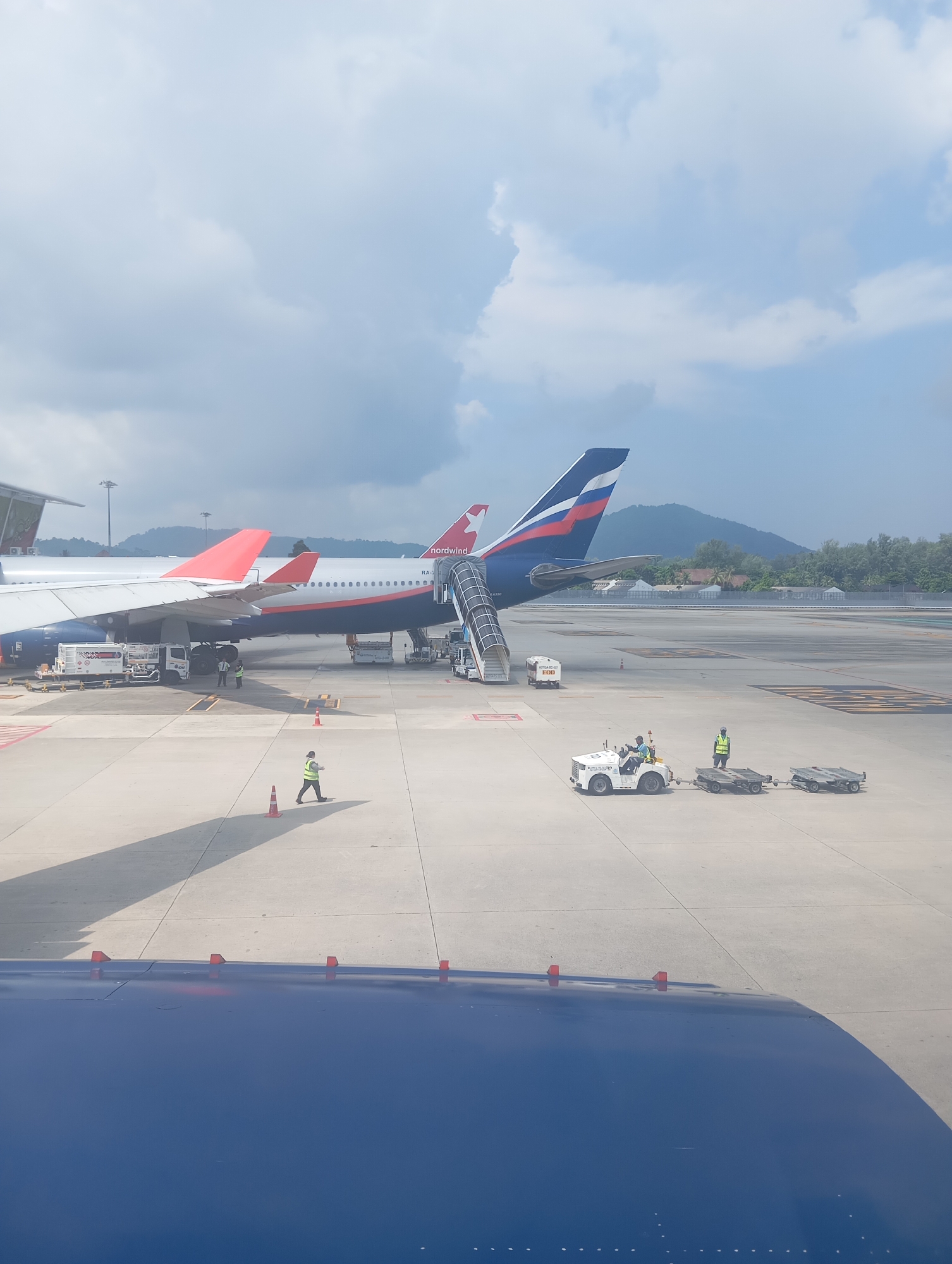 The image captures an airport tarmac scene in Tambon Mai Khao, Thailand, under a partly cloudy sky. Two airplanes are visible: a white and red-tailed aircraft on the left and a blue and red-tailed aircraft, branded "nordwind", on the right, with a jet bridge attached. Several airport vehicles and personnel are present. A white tug vehicle, driven by an individual in a green vest, is towing two flatbed carts. Another person in a green vest is standing near the tug. Two other individuals in green vests are standing near the base of the blue and red-tailed airplane, possibly performing ground operations. The ground is paved concrete, marked with yellow lines. In the foreground, the edge of a blue aircraft wing is visible. Distant trees and mountains form the background.