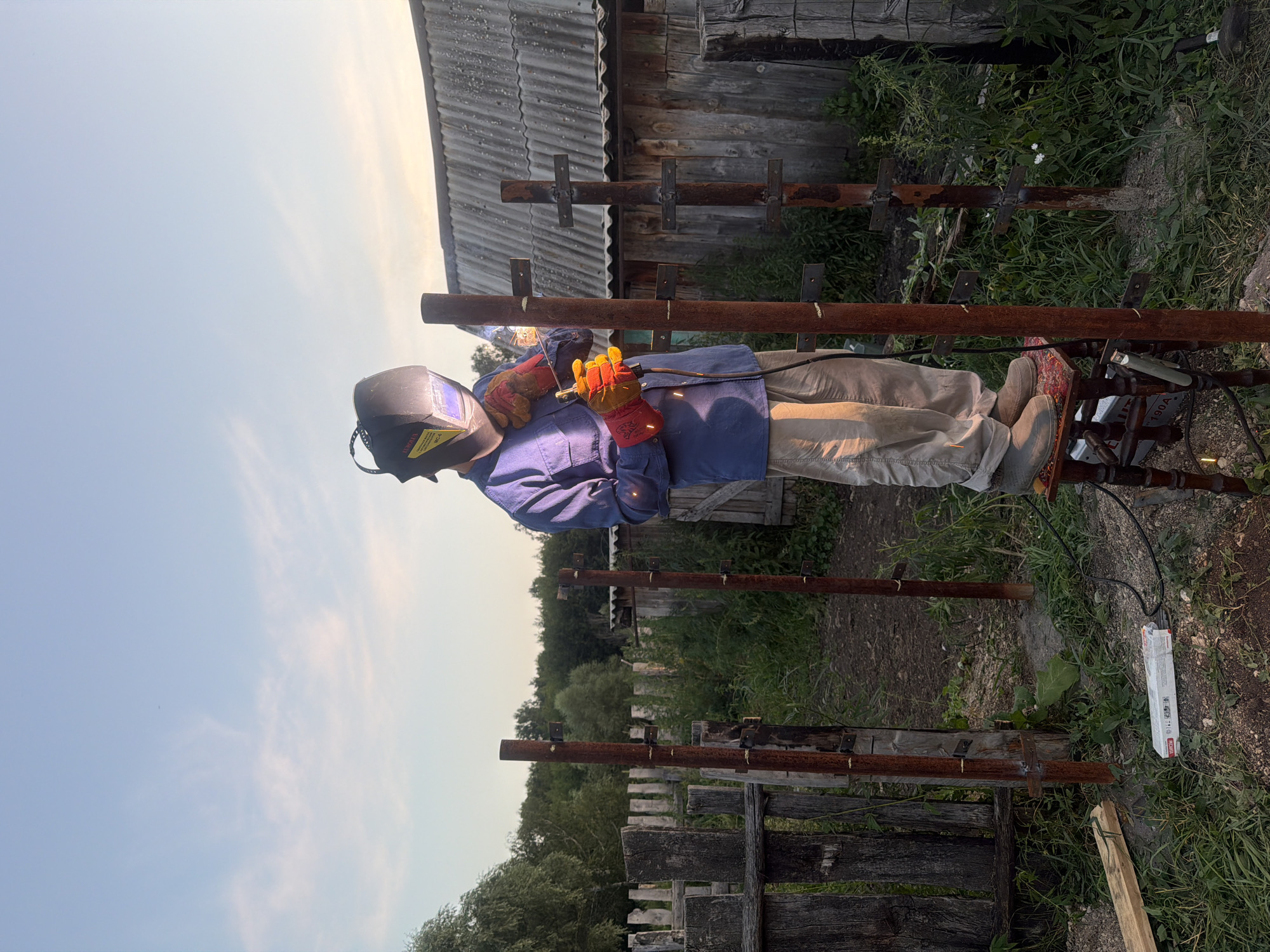 A man wearing a blue shirt, tan pants, a welding mask, and orange gloves stands on a red and black stool welding two rusty metal posts. The welding equipment is connected to a power source visible on the ground. The scene is outdoors, with a wooden fence and a rustic wooden building in the background. Trees and overgrown grass surround the area. The sky is blue with some scattered clouds. This activity appears to be taking place in the rural settlement of Kuseevo, Russia, likely in a backyard or work area.