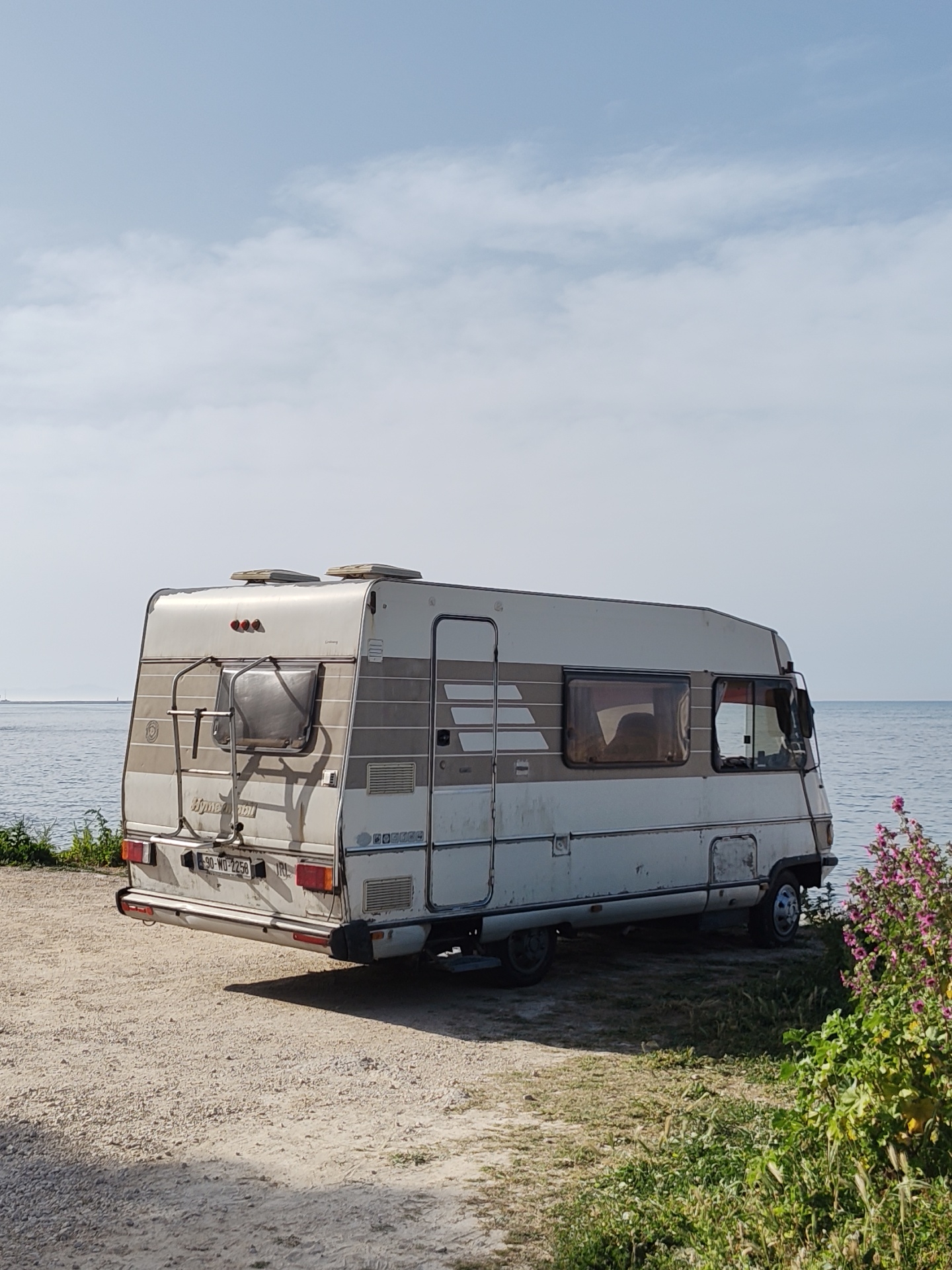 A Hymer campervan is parked on a dirt lot bordering the sea in Dénia, Spain. The van, predominantly white with brown stripes, faces the viewer, its rear featuring a ladder and a partially obscured window. The license plate reads "90-WD-2256 IRL." The sea is visible in the background, meeting a light blue, cloud-dotted sky. Vegetation, including flowering bushes, grows to the right and left of the van. The ground is dirt and gravel with patches of grass.