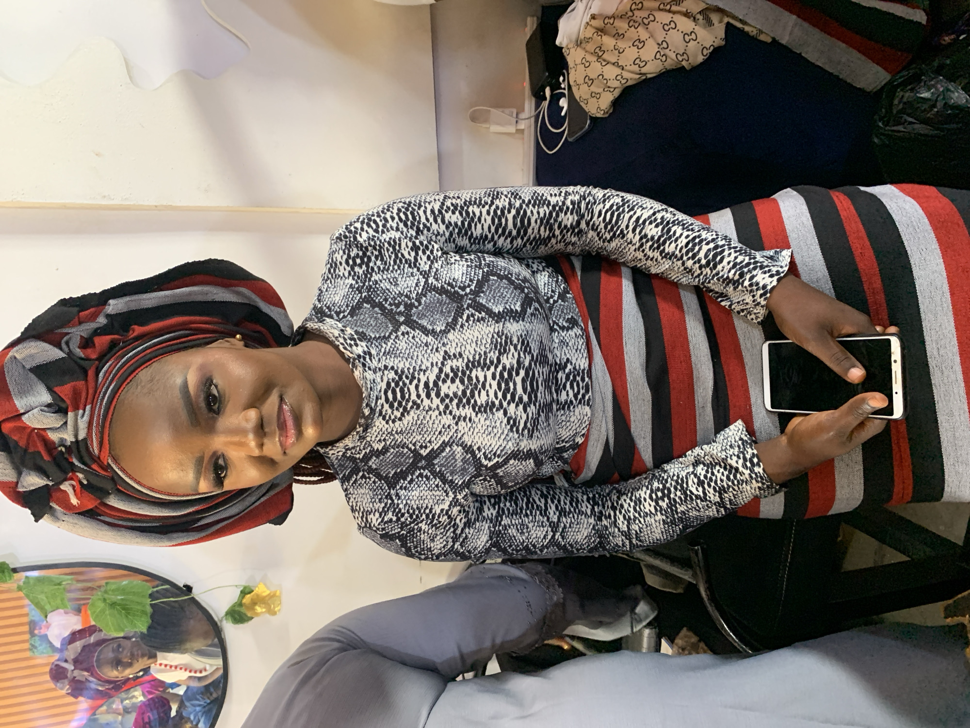 A woman sits indoors, holding a smartphone in her right hand. She wears a striped headwrap, a snake-print top, and a striped skirt or dress. She appears to be seated in a chair. The background includes other items of clothing and a room with white walls. The image does not contain enough information to confirm this location in Jos, Nigeria.