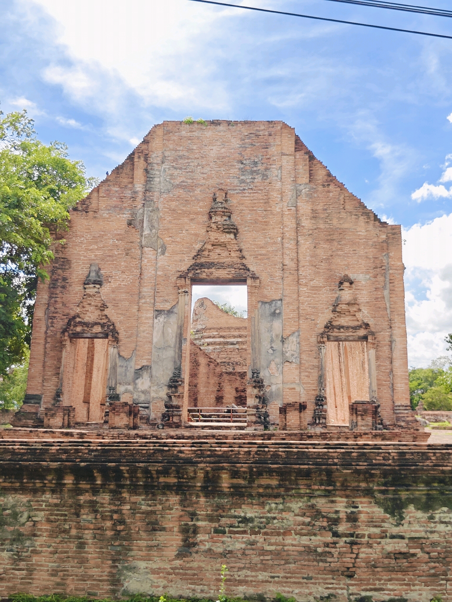 A full-frame, eye-level shot captures the weathered facade of an ancient brick temple ruin under a bright, cloudy sky in Ayutthaya, Thailand. The temple's central, arched opening reveals a glimpse of another crumbling structure within. Flanking the central opening are two recessed alcoves, each adorned with decorative brickwork and a hint of faded fabric. The upper edges of the temple are jagged, showing signs of decay and erosion. Lush green trees frame the left side of the structure, and the foreground is dominated by a lower brick wall. The sky is a vivid blue, punctuated by fluffy white clouds. Power lines are visible near the top of the frame.